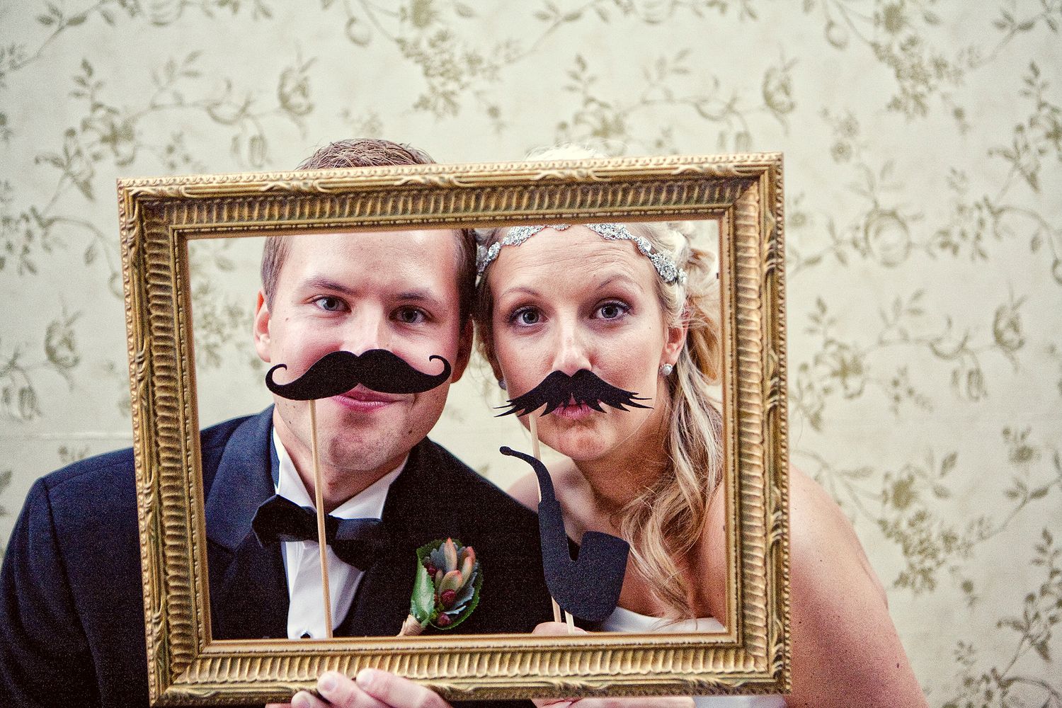 Bride and groom pose with fake mustaches inside a gold picture frame at wedding photo booth.