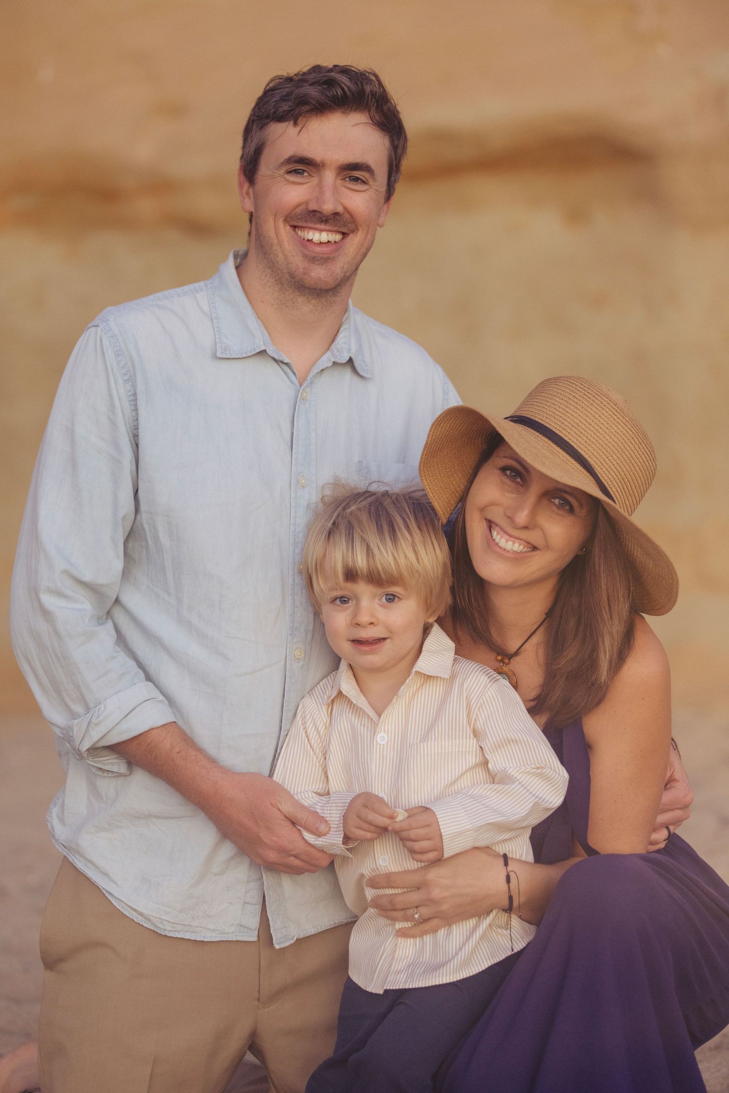 Happy family portrait of parents and young son posing outdoors near sandy rock formation.