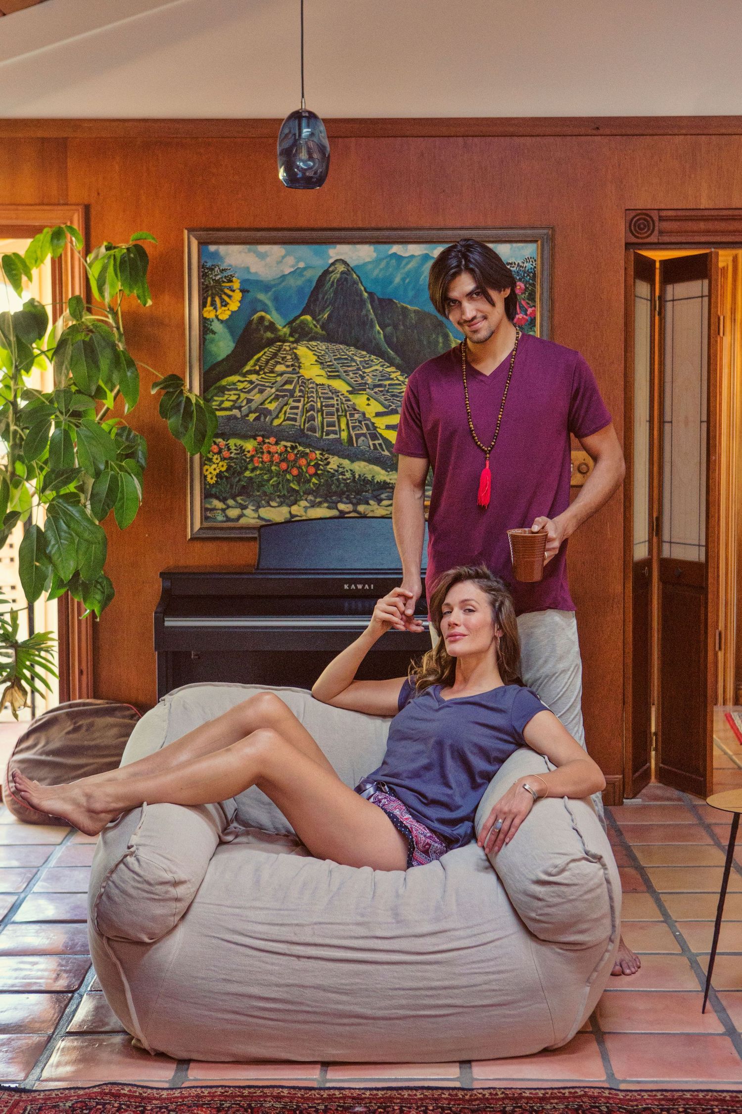 Couple relaxing at home with Machu Picchu painting, woman on bean bag chair, man standing holding mug.
