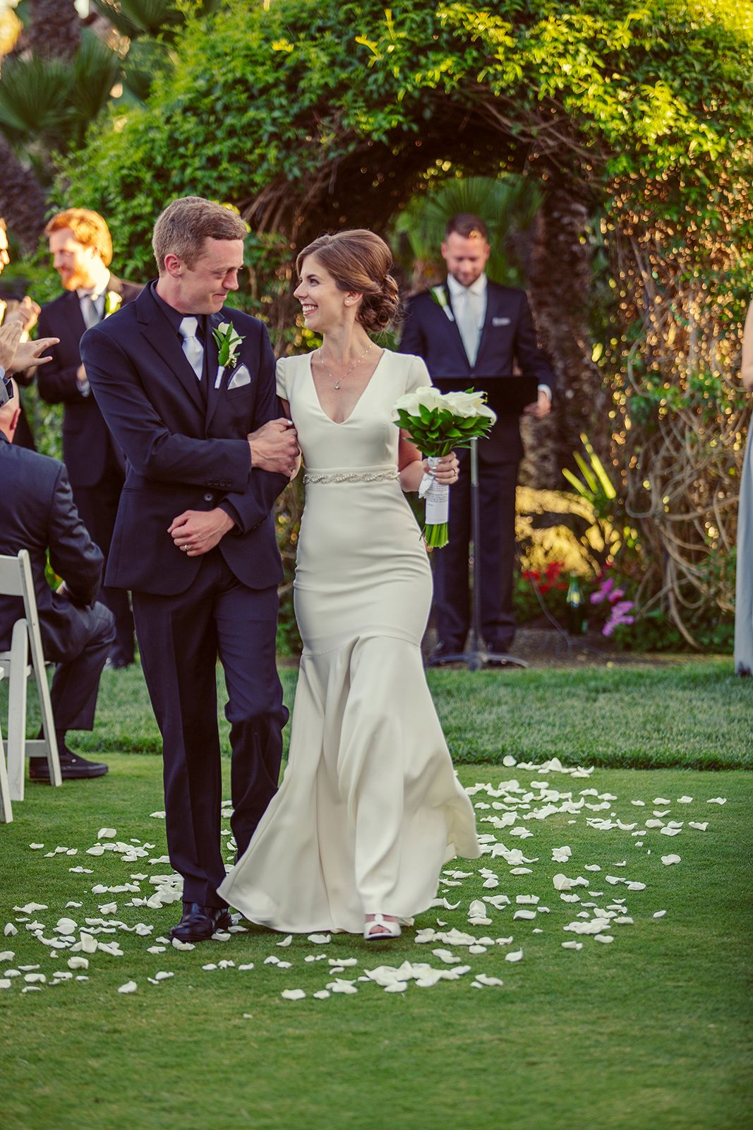 Smiling bride in elegant mermaid gown walks down petal-lined aisle with groom in navy suit at garden wedding.