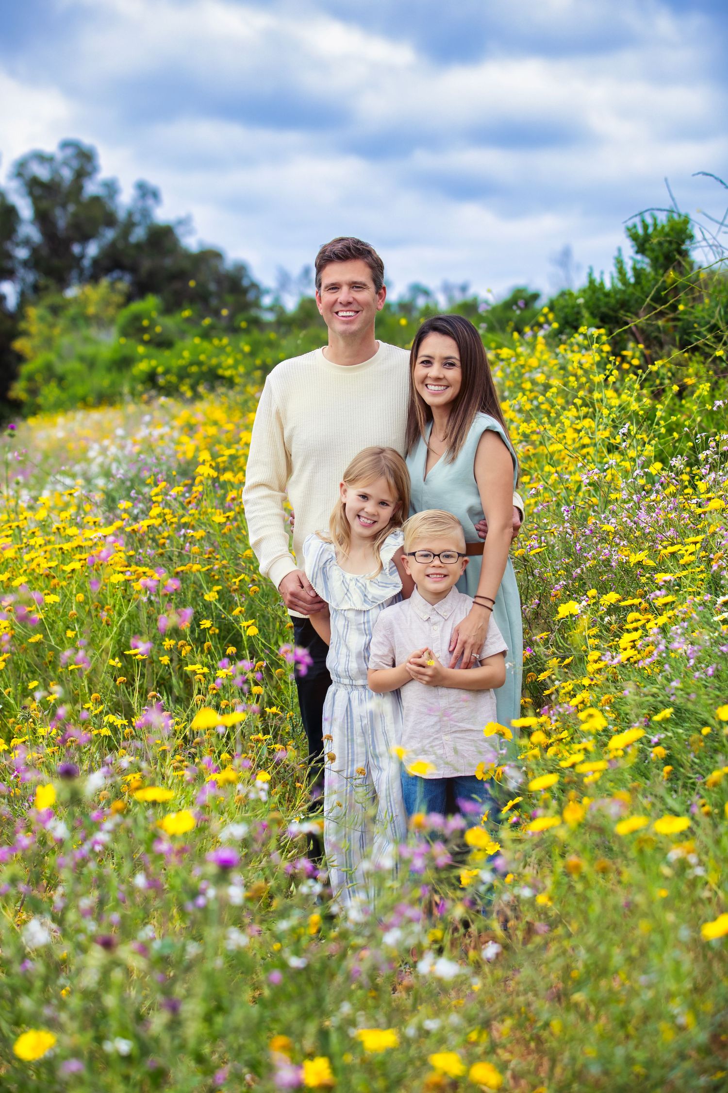 Happy family of four posing in a vibrant wildflower meadow with yellow blooms under a partly cloudy sky.