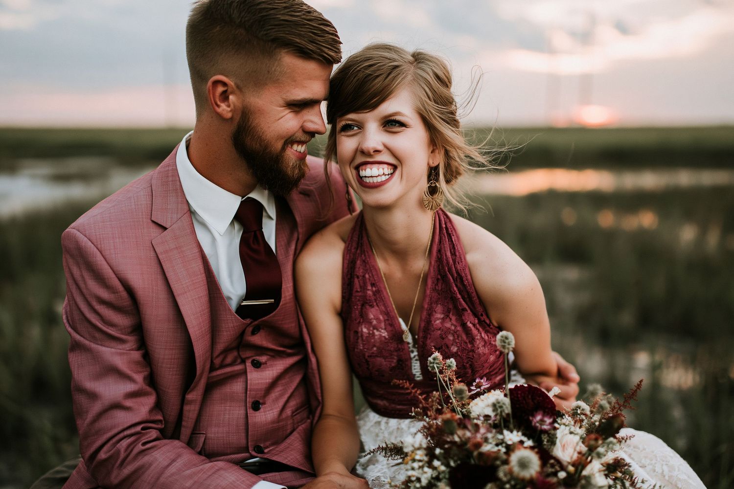 A smiling bride holding a burgundy bouquet sits with her groom in a pink suit at sunset near a marsh.