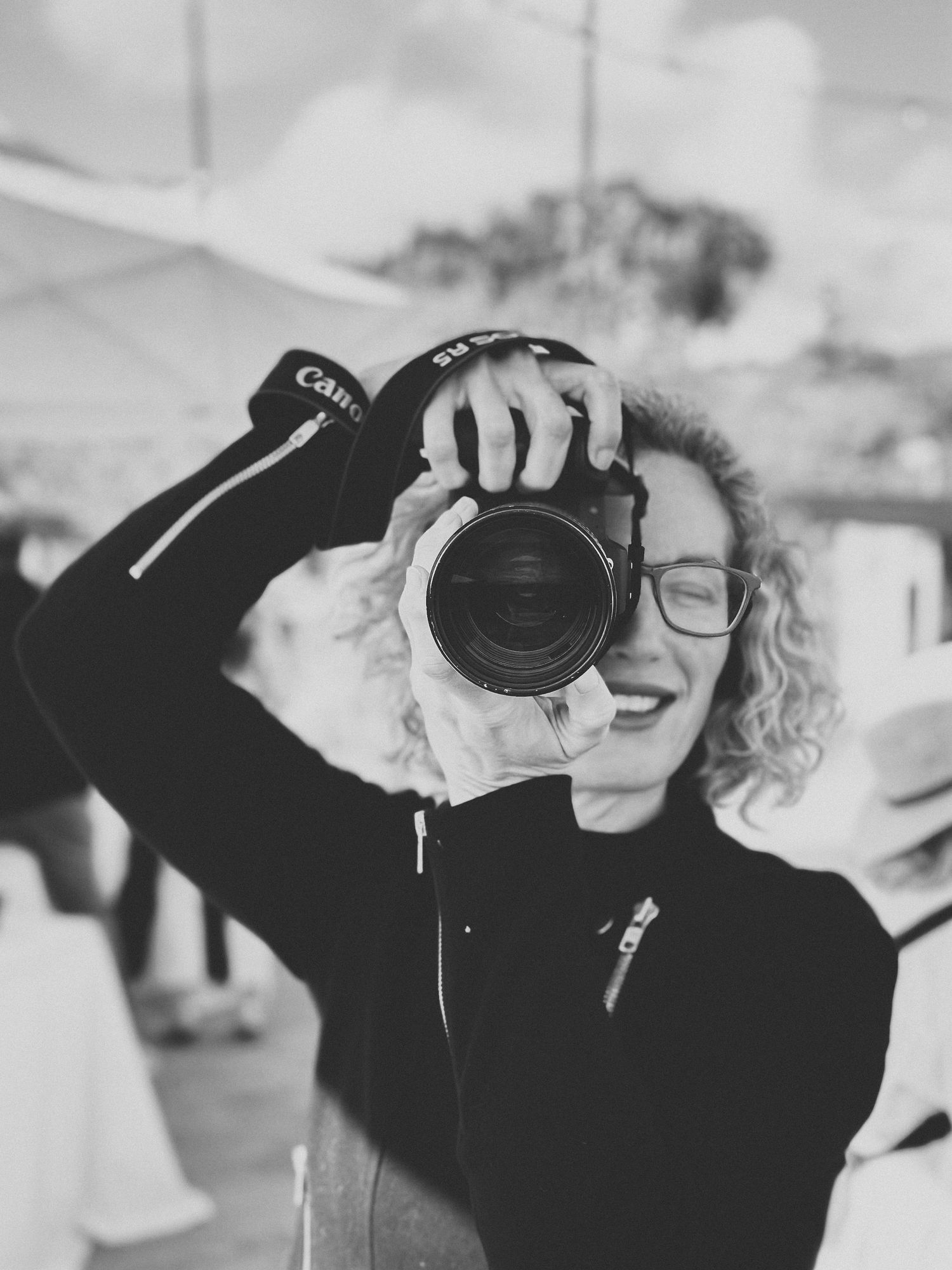 Black and white photo of a smiling woman with curly hair holding a Canon camera up to photograph.