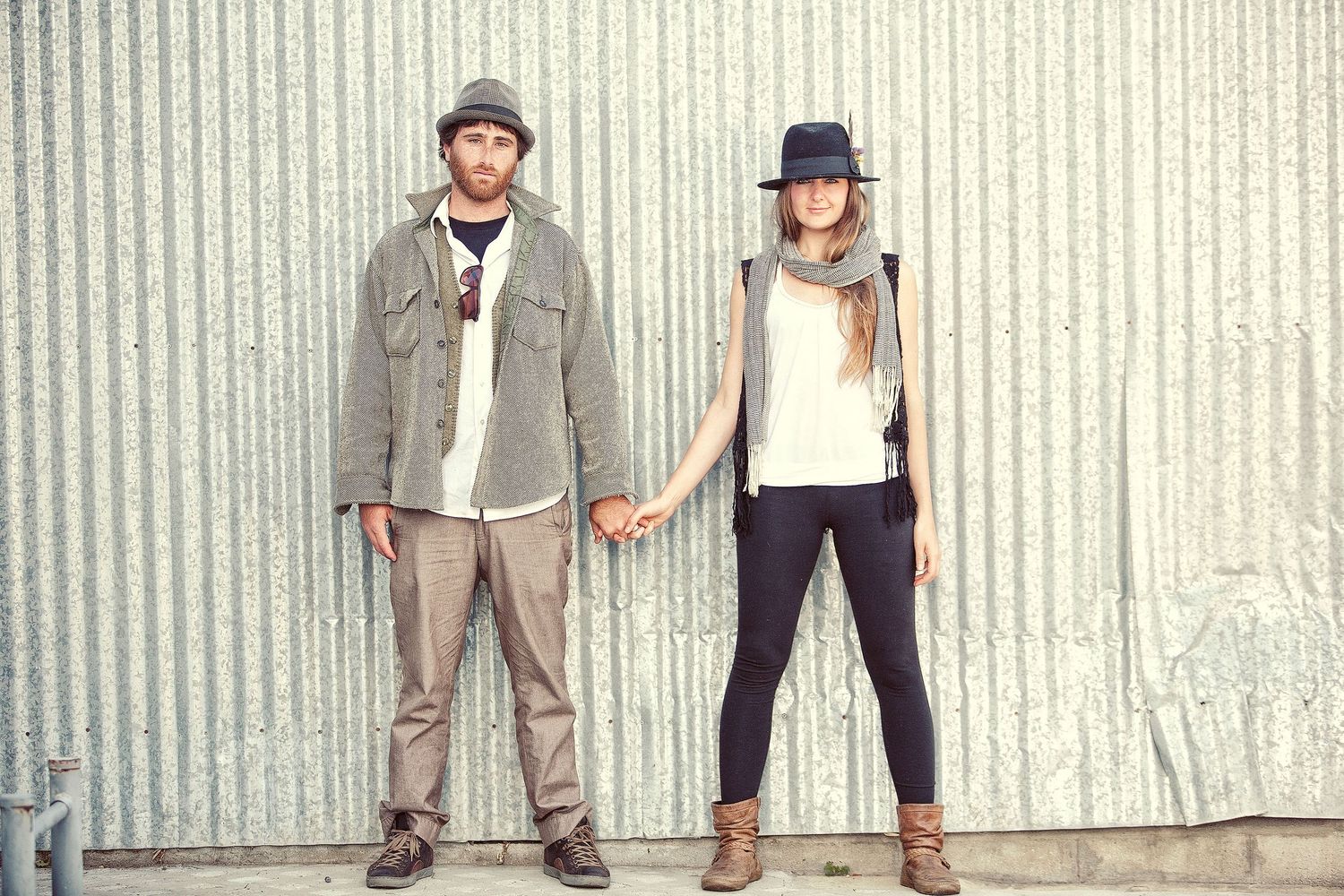 A stylish couple holding hands in front of a corrugated metal wall, both wearing hats and boots in casual outfits.