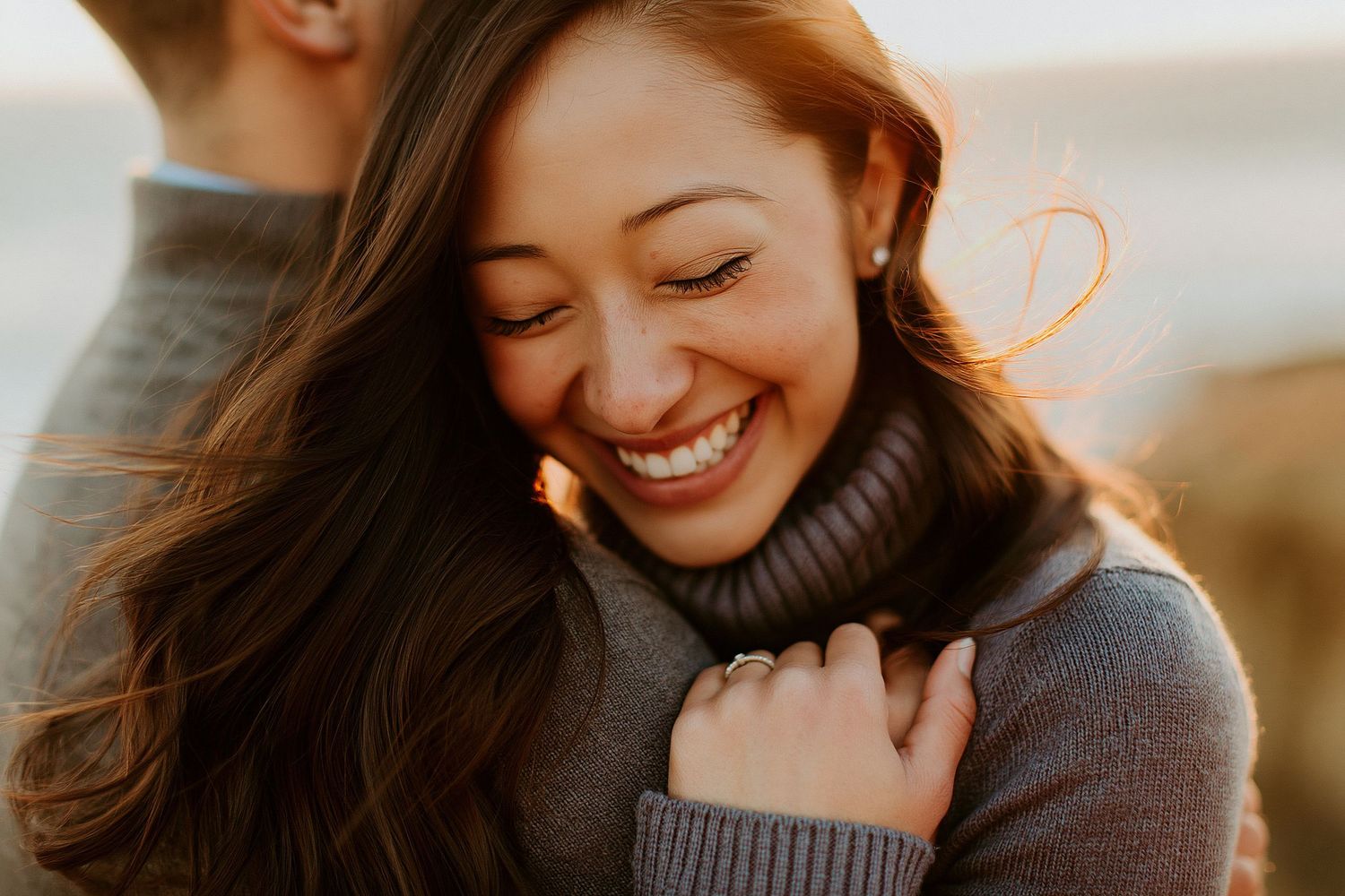 A smiling woman with long brown hair hugs a man outdoors, wearing a turtleneck sweater and engagement ring.