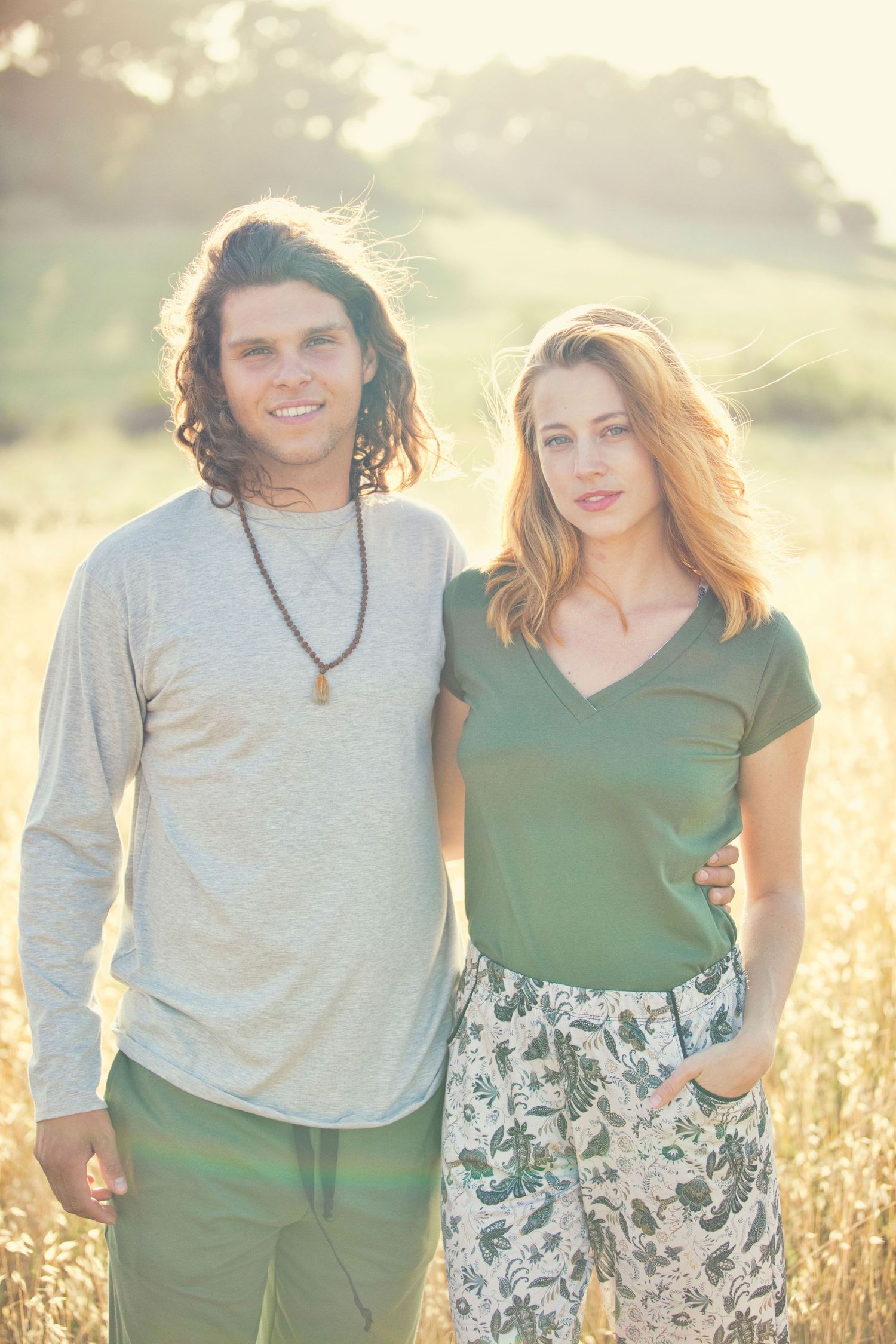 Young couple standing in golden sunlit field, man in gray shirt with beads, woman in green top and floral pants.