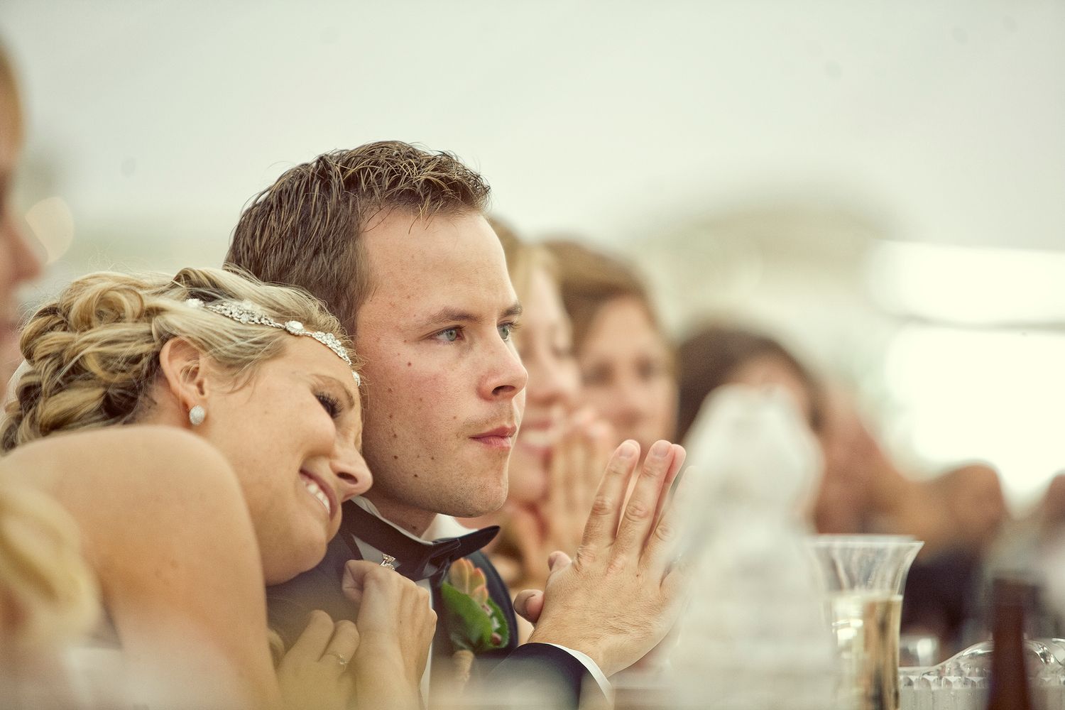 Bride and groom share a tender moment at their wedding reception, smiling and listening to speeches.