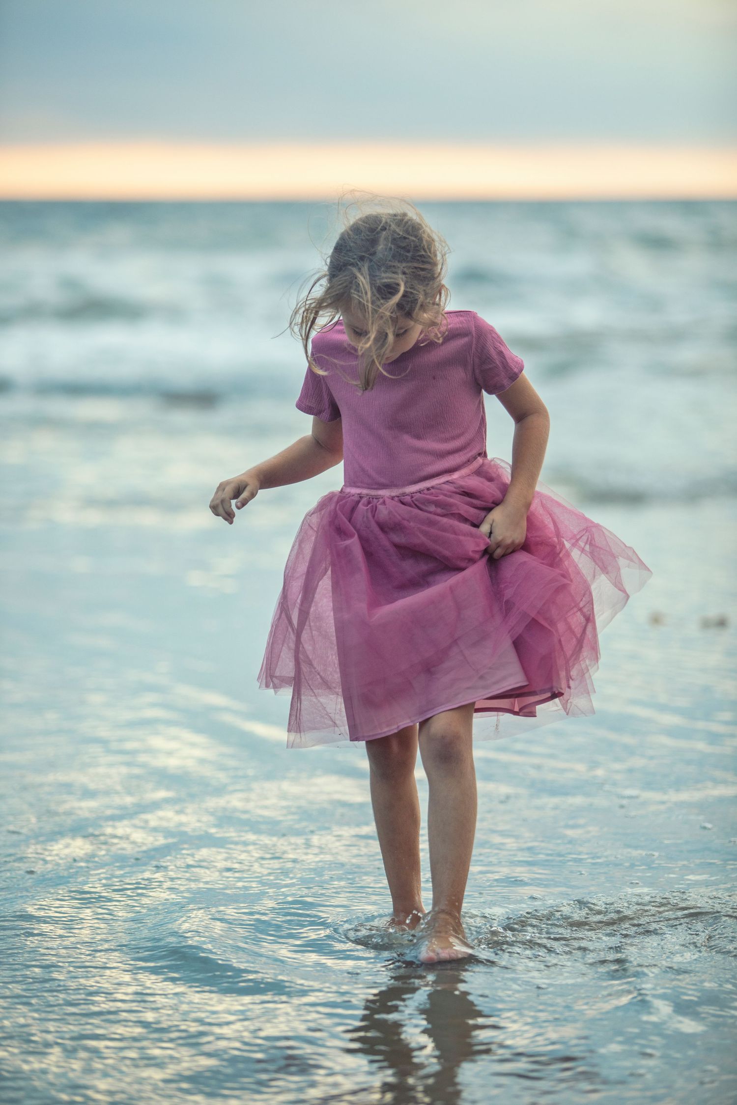 Young girl in pink tutu dress wading barefoot at ocean shore during sunset, viewed from behind.