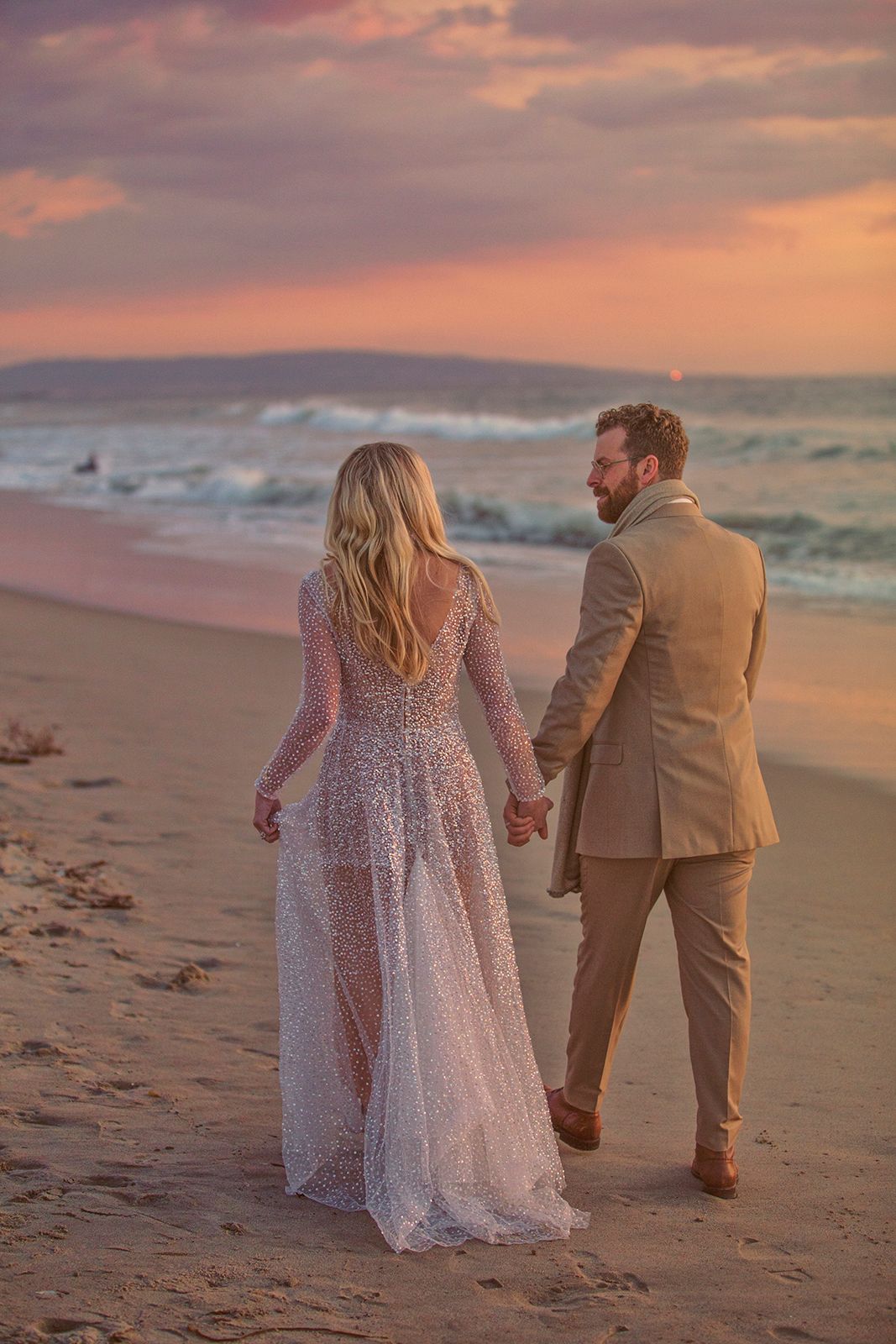 Couple in wedding attire holding hands walking on a sandy beach at sunset with colorful pink sky.