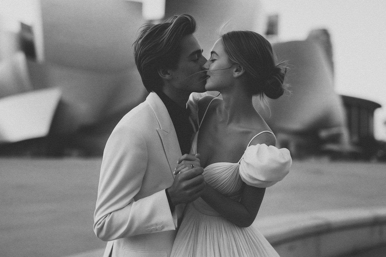 Black and white photo of a couple sharing a romantic kiss outdoors, dressed in elegant wedding attire.