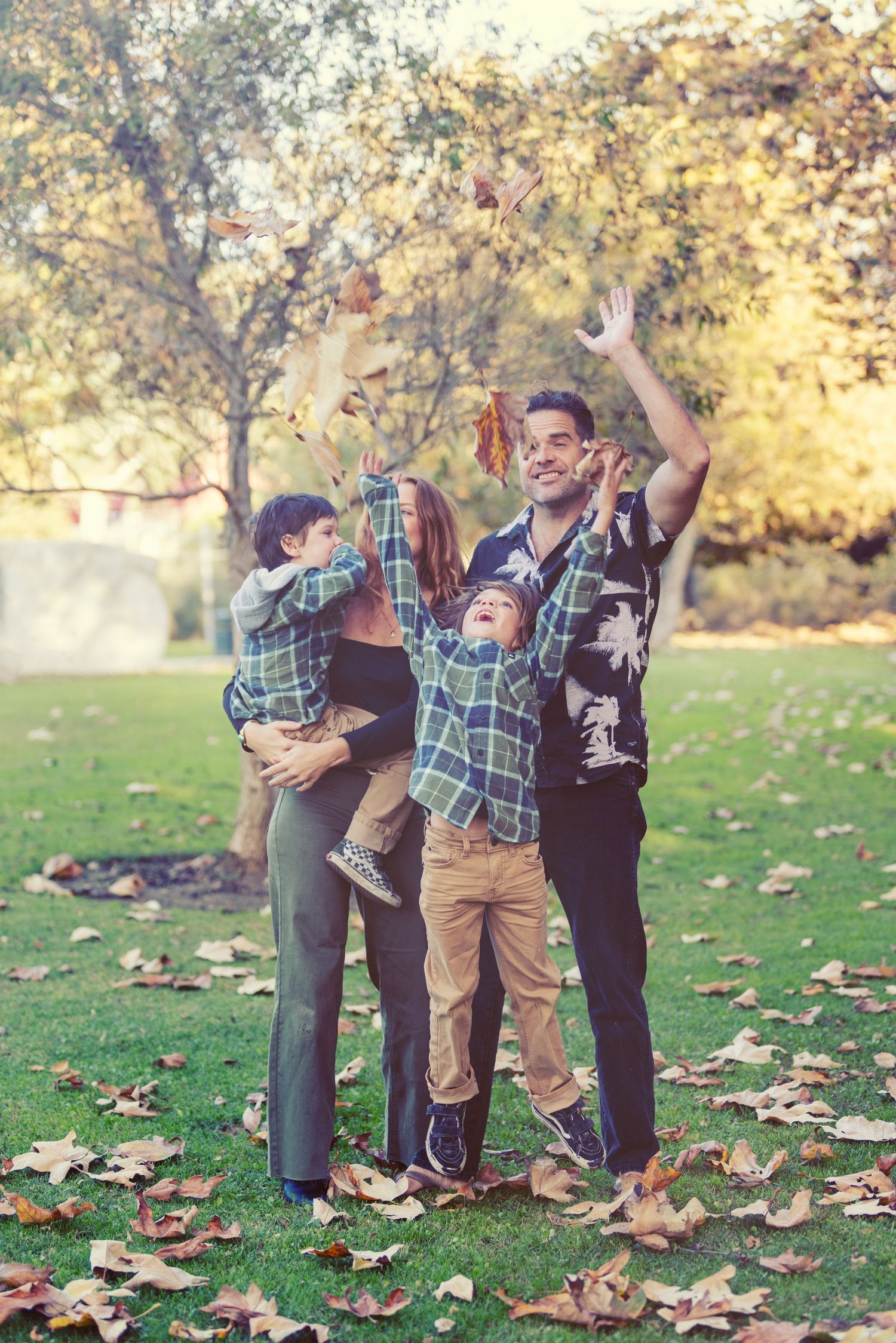 Family joyfully tossing autumn leaves in a park, with two children and parents smiling outdoors in fall foliage.