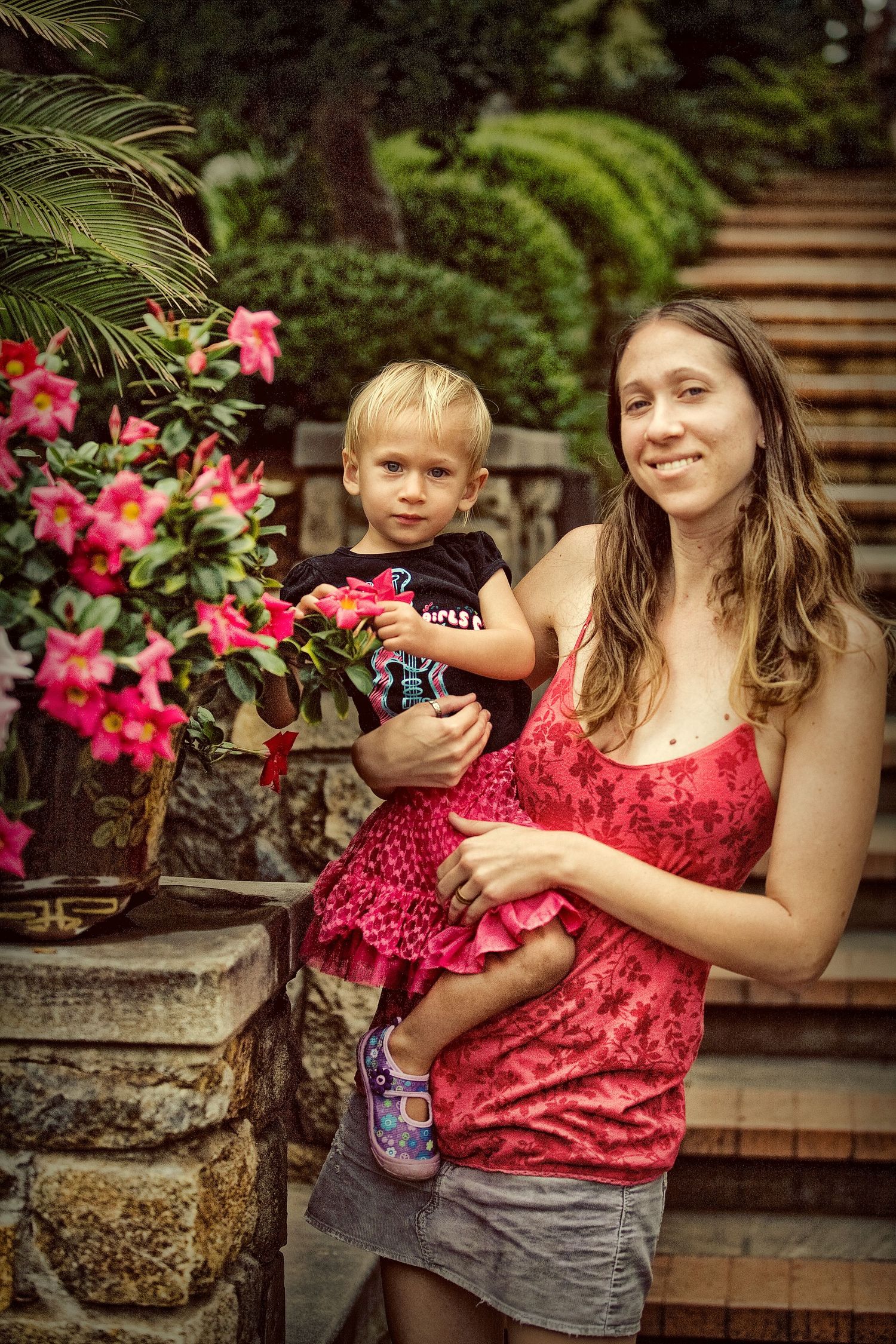 A smiling woman in a red top holds a young blonde toddler near vibrant pink flowers in a lush garden setting.