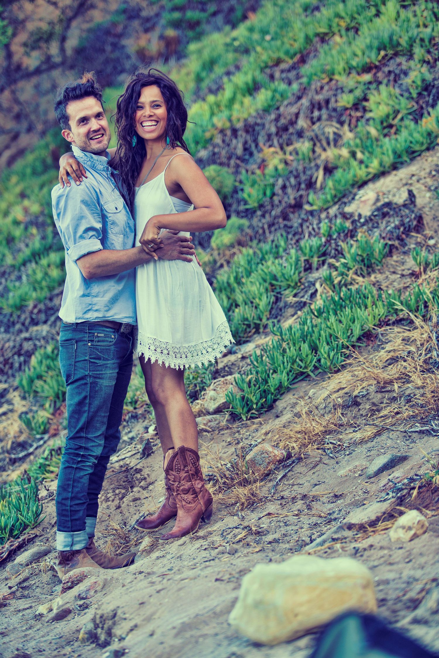 Happy couple embracing outdoors on a rocky hillside, woman in white dress and cowboy boots, man in denim shirt.