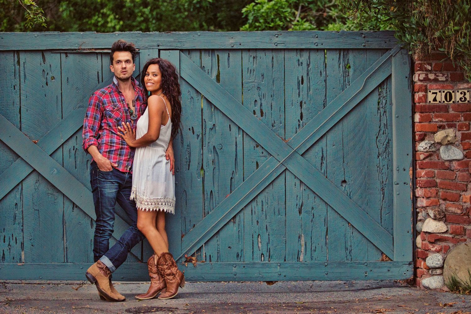 A stylish couple poses against a rustic teal wooden gate, wearing casual country-chic outfits with cowboy boots.