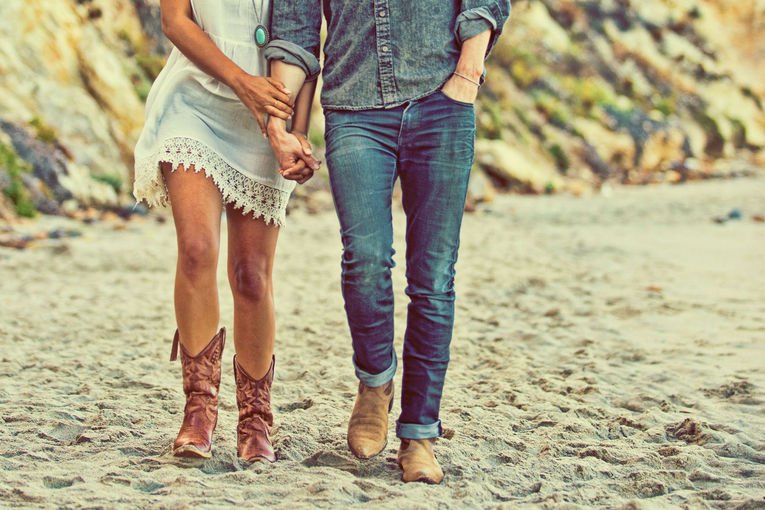 Couple holding hands on sandy beach, woman in white dress and cowboy boots, man in jeans and denim shirt.