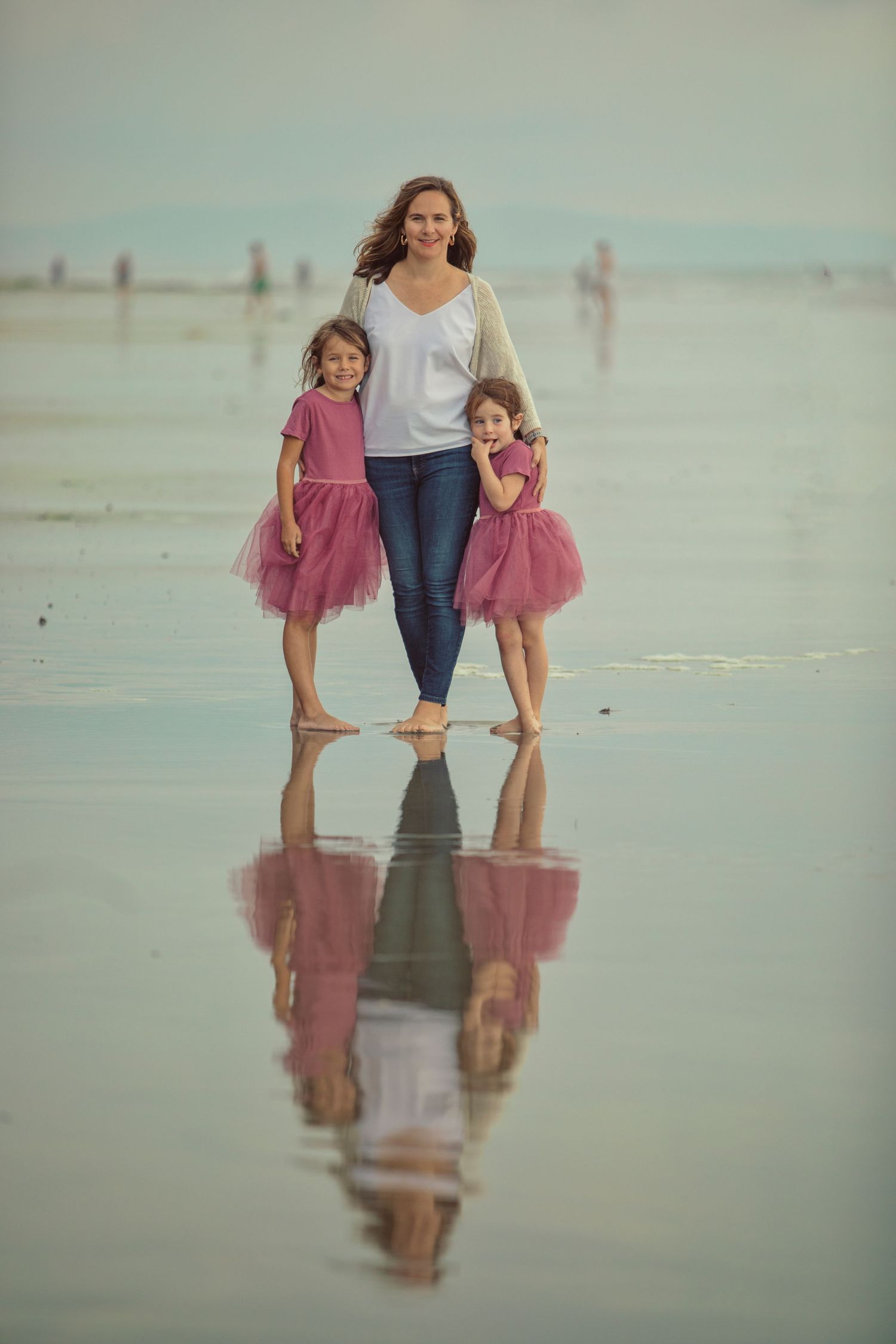 A mother and two young girls in pink tutu skirts walk barefoot on a reflective beach shoreline.