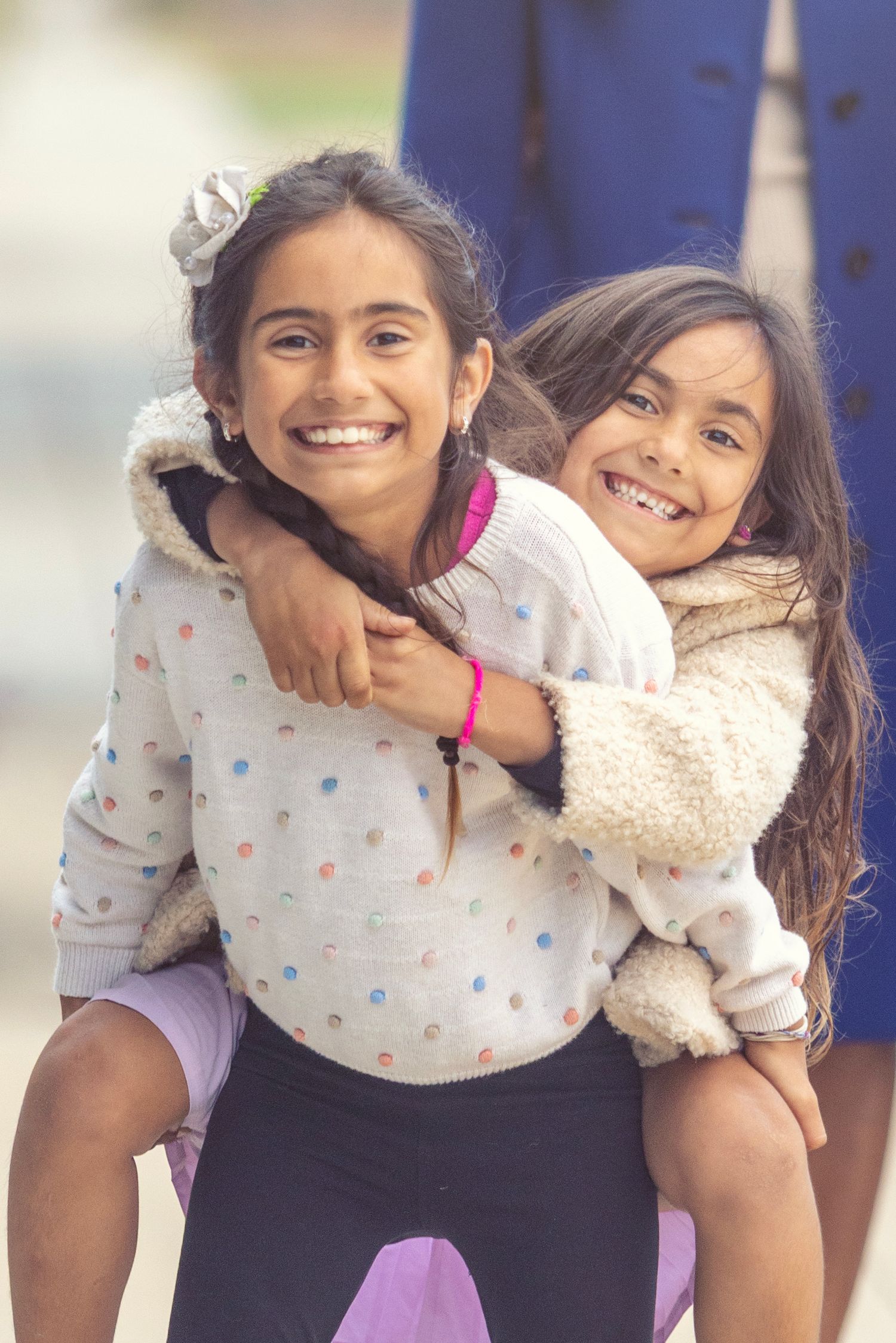 Two young girls smiling joyfully, one giving the other a piggyback ride outdoors in cozy sweaters.