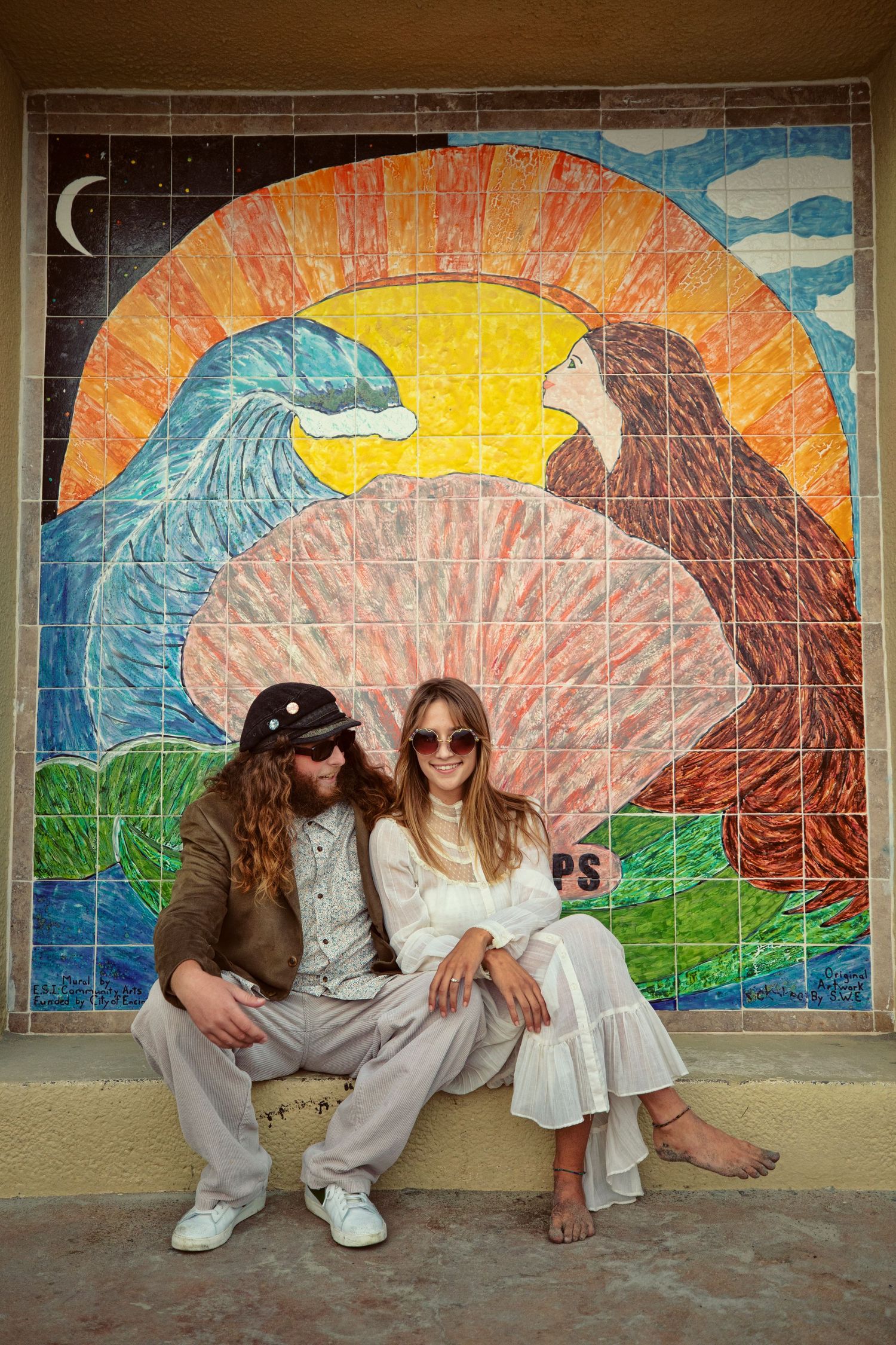 Couple sitting in front of a colorful mosaic tile mural featuring waves and a sunset in retro boho style.