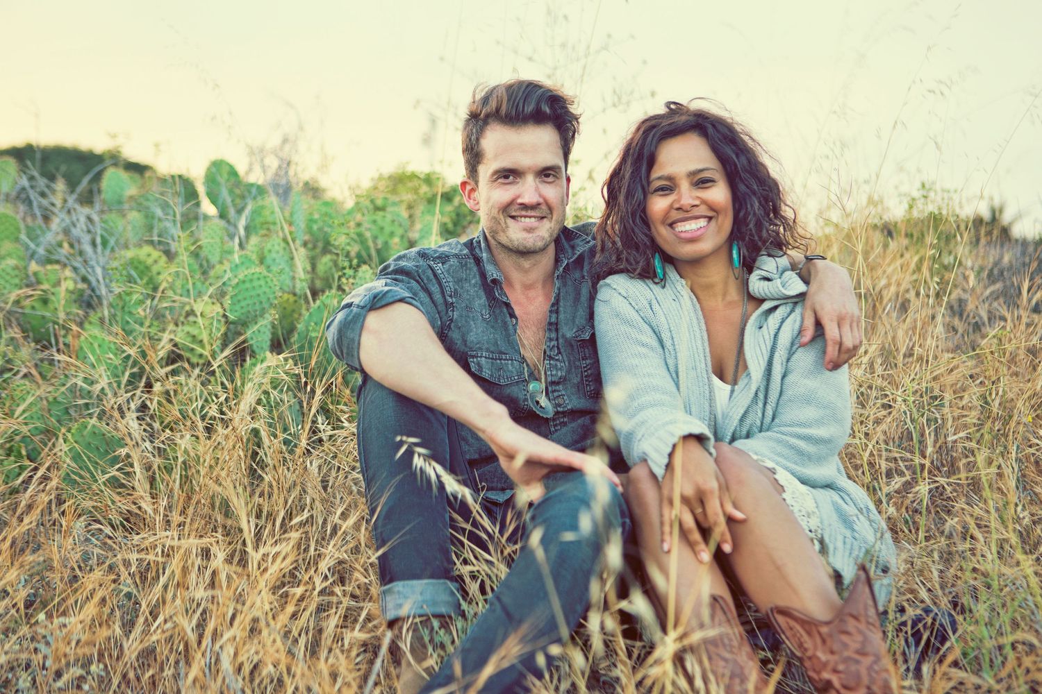 Couple sitting in tall grass field with cactus, smiling and embracing in a warm outdoor setting.