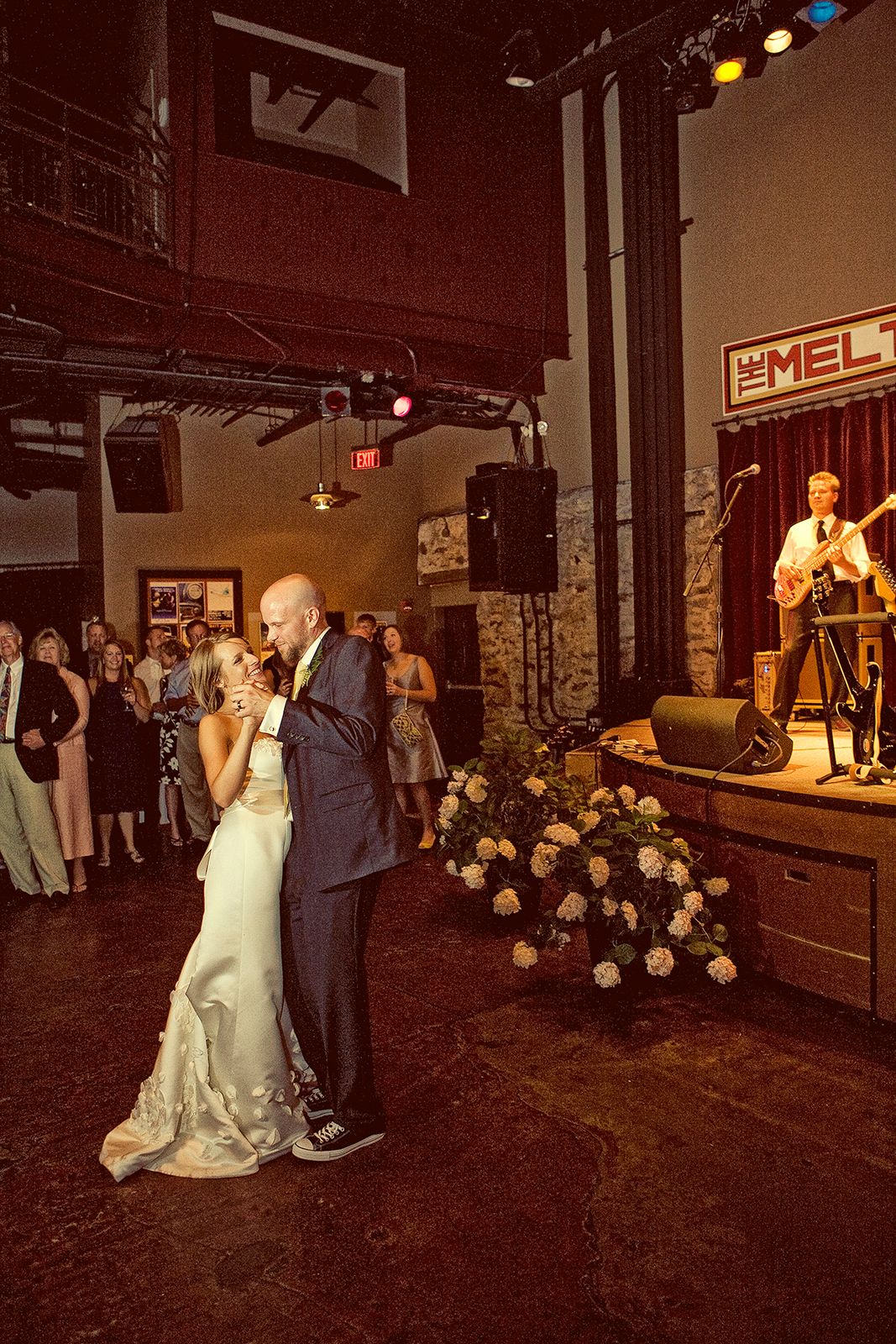 Bride and groom share first dance at wedding reception venue with live band performing on stage in background.