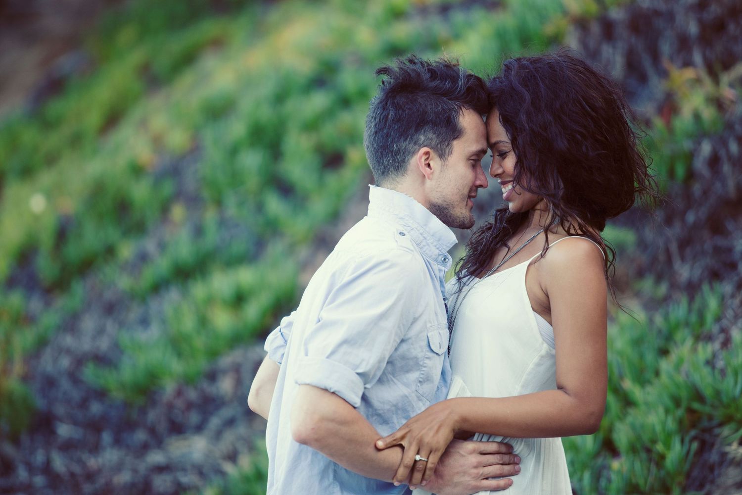 A romantic couple embracing outdoors, sharing an intimate moment with lush green hillside in the background.