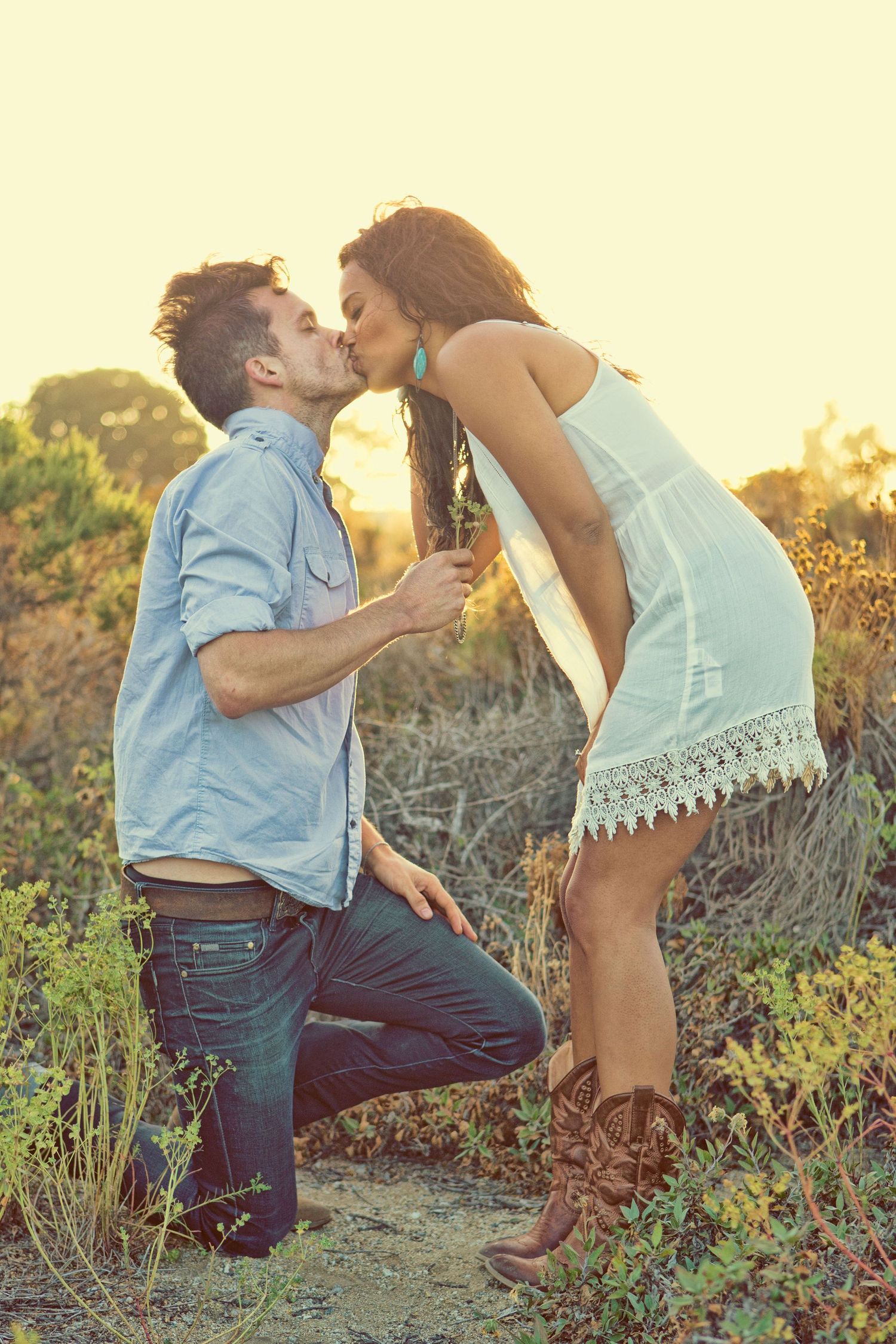 Couple sharing romantic kiss during outdoor proposal at golden hour in natural setting.