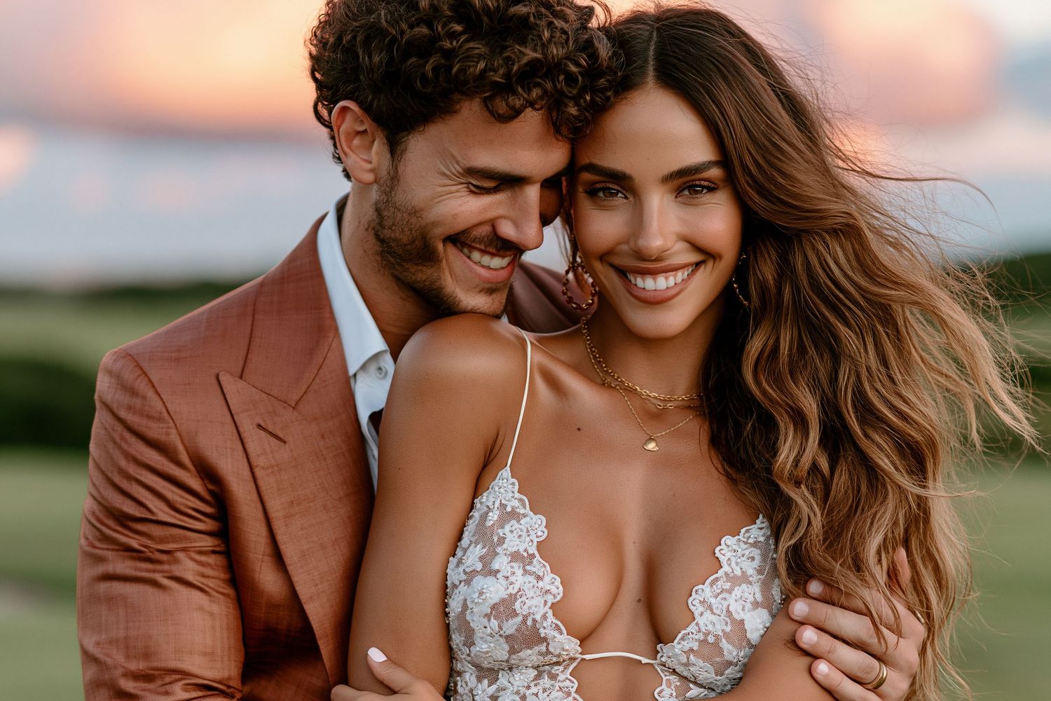A smiling couple in wedding attire embraces outdoors at sunset, man in tan suit, woman in lace bridal gown.