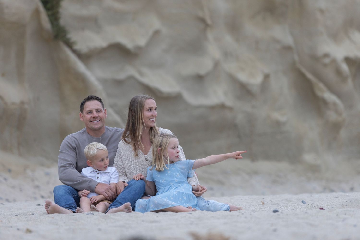 Family of four sitting on sandy beach with rocky cliffs, two parents and two young children pointing at the ocean.