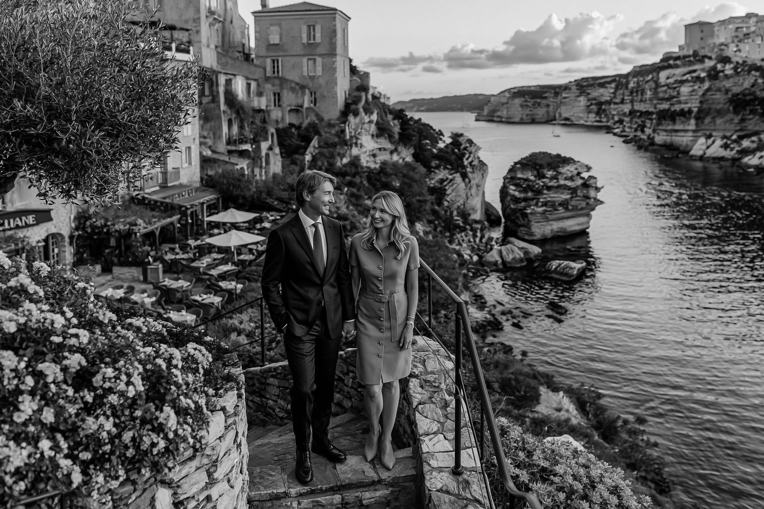 Black and white photo of a couple walking along scenic coastal stone steps beside a Mediterranean village.