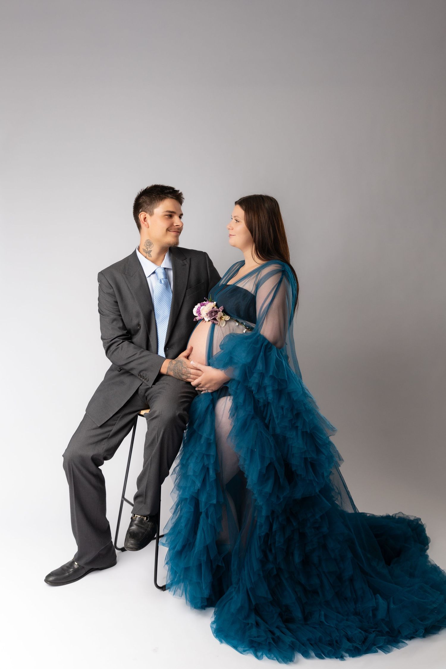 A couple in formal attire poses together against a gray backdrop, with the woman wearing a flowing blue tulle gown.