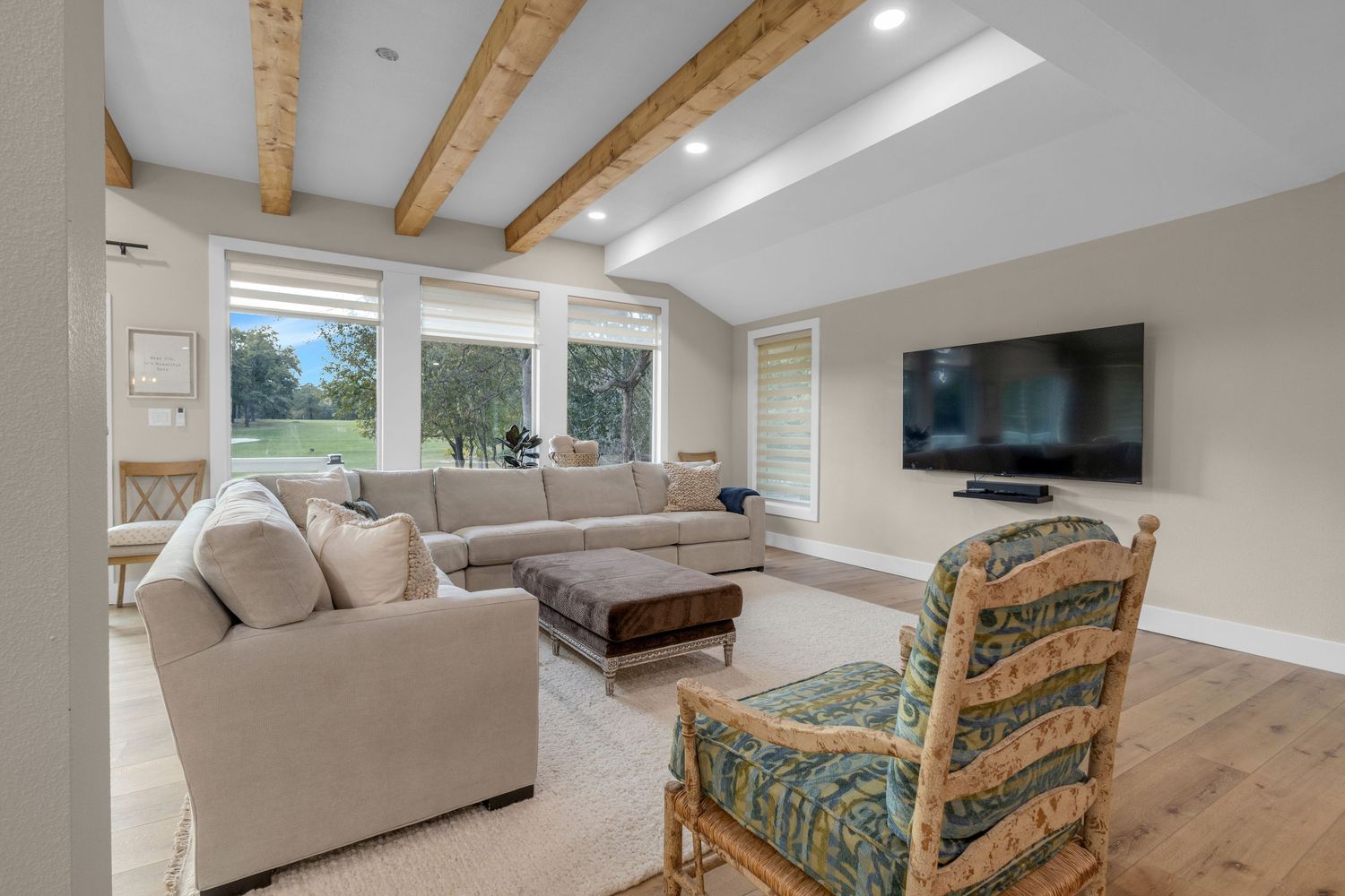 Modern living room with exposed wood beams, neutral sectional sofa, patterned accent chair and wall-mounted TV.