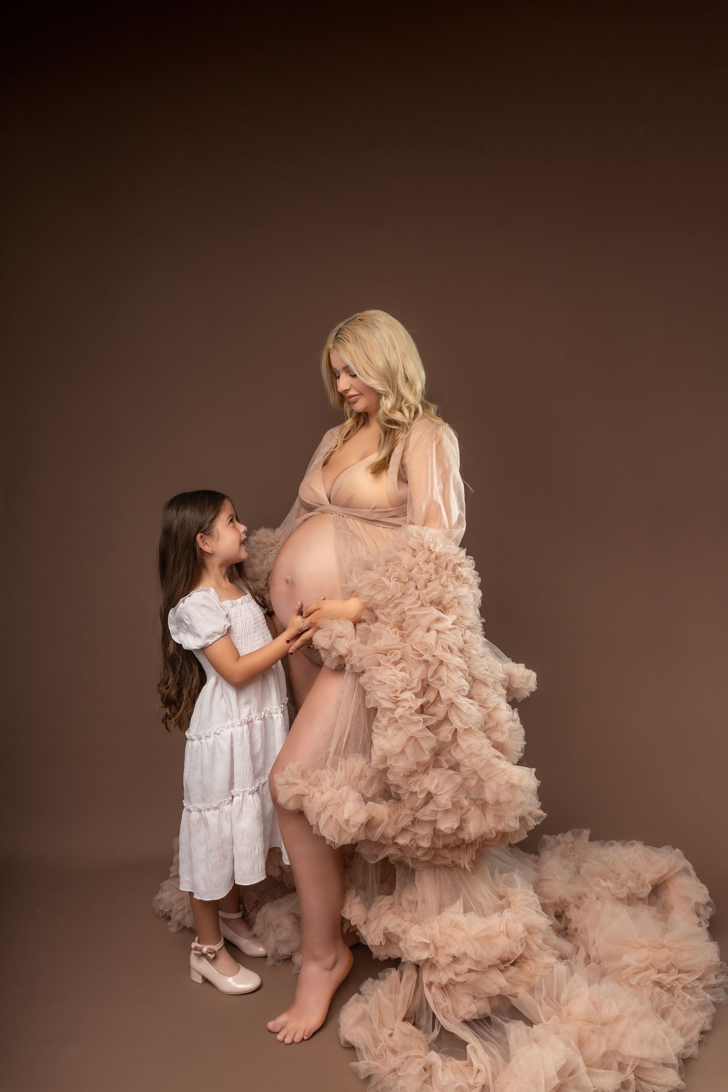 A child in white dress interacts with someone wearing a flowing blush-colored tulle gown in a studio setting.