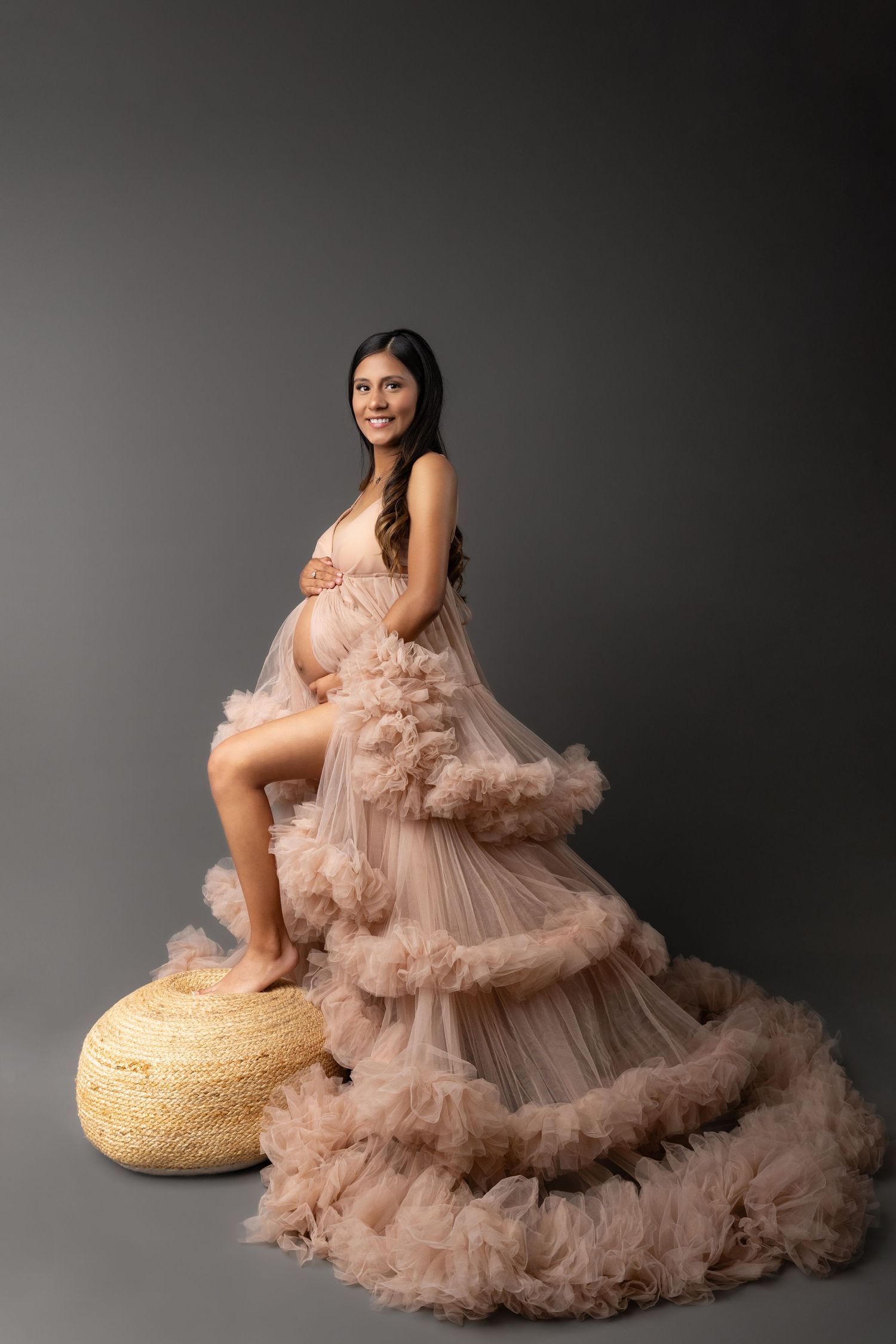 Elegant maternity portrait featuring a flowing blush tulle gown with ruffled tiers and a rustic woven footstool against gray backdrop.