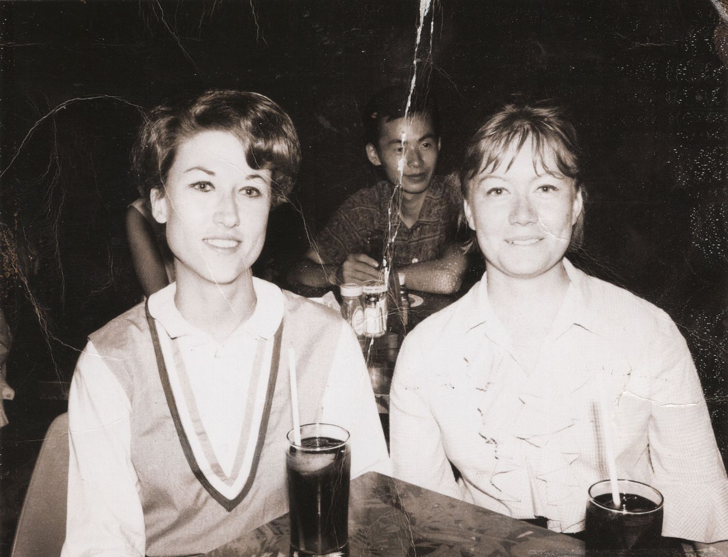 Two women sitting at a table with dark drinks in a vintage black and white photo, man visible in background.