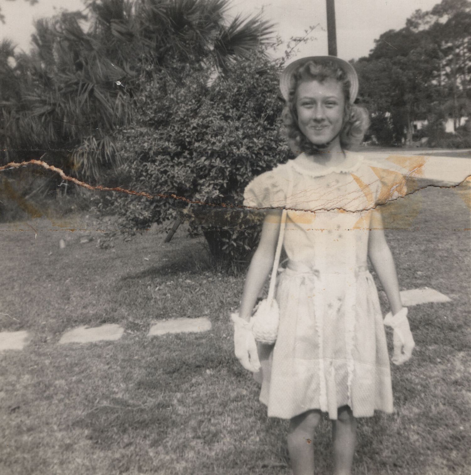 Vintage black and white photo of a young girl in a white dress and hat posing outdoors near tropical plants.