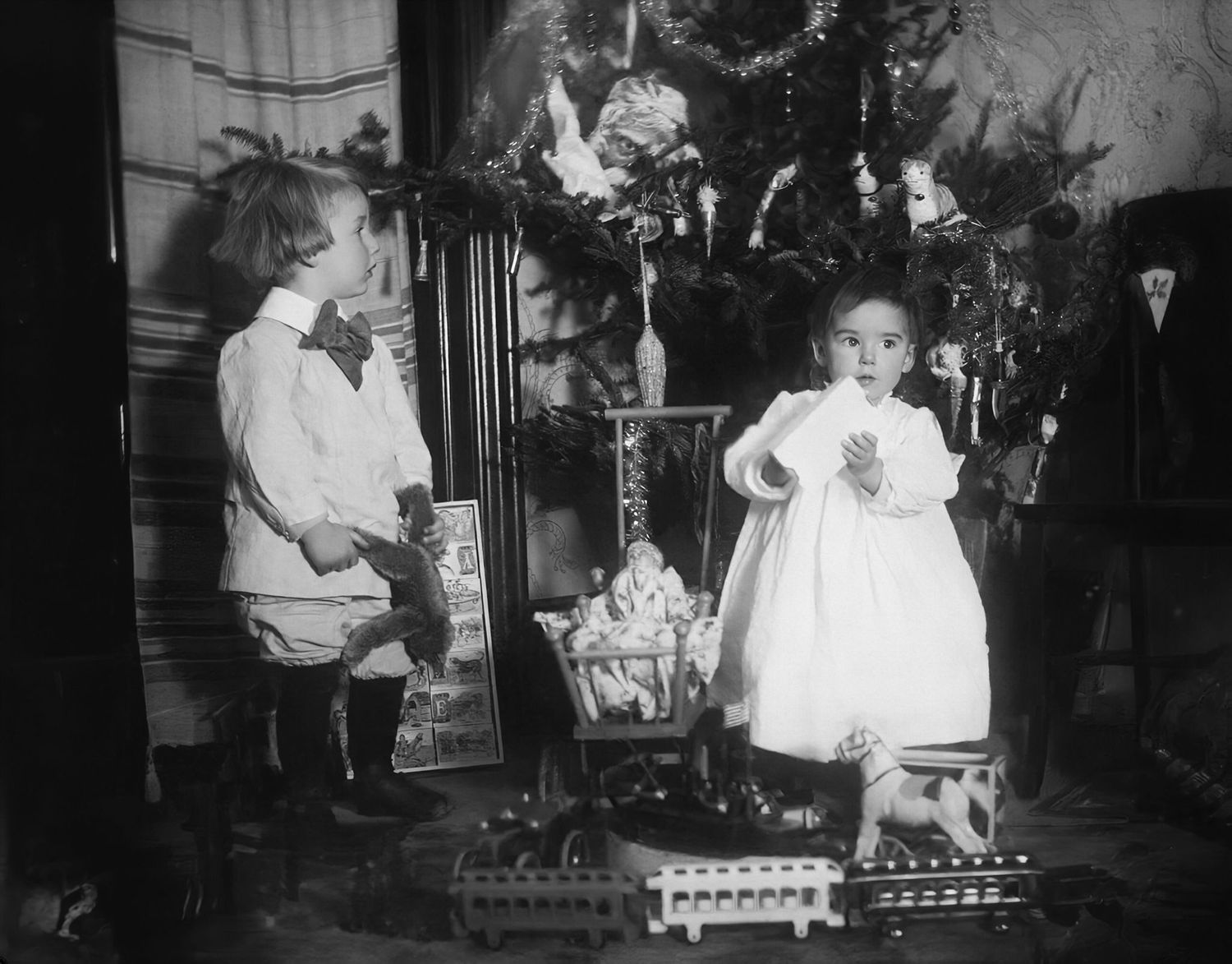 Vintage black and white photo of two children standing by a decorated Christmas tree with toys and gifts.