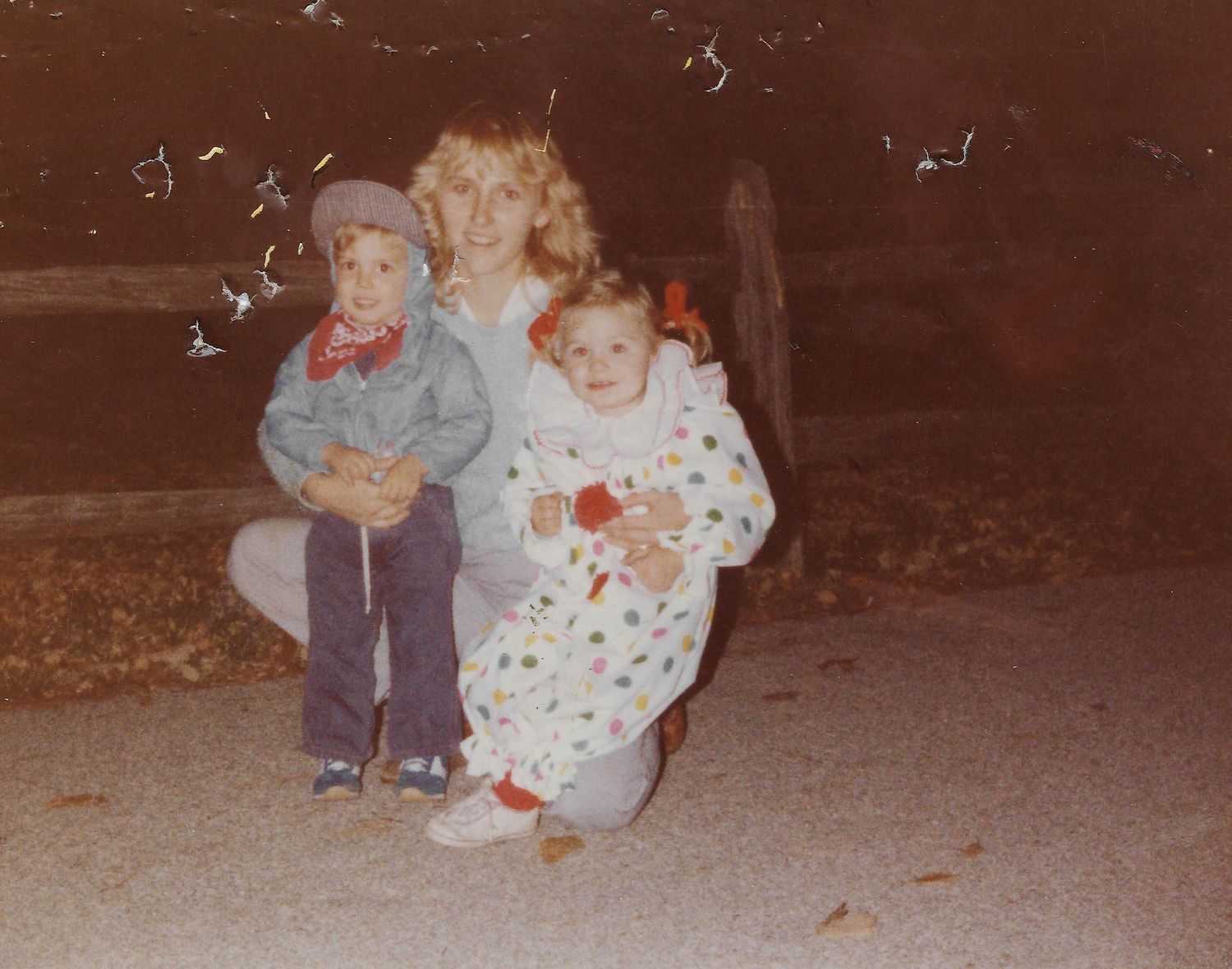 A woman crouches with two young children in Halloween costumes, a clown and a cowboy, outdoors at night in a vintage 1970s photo.