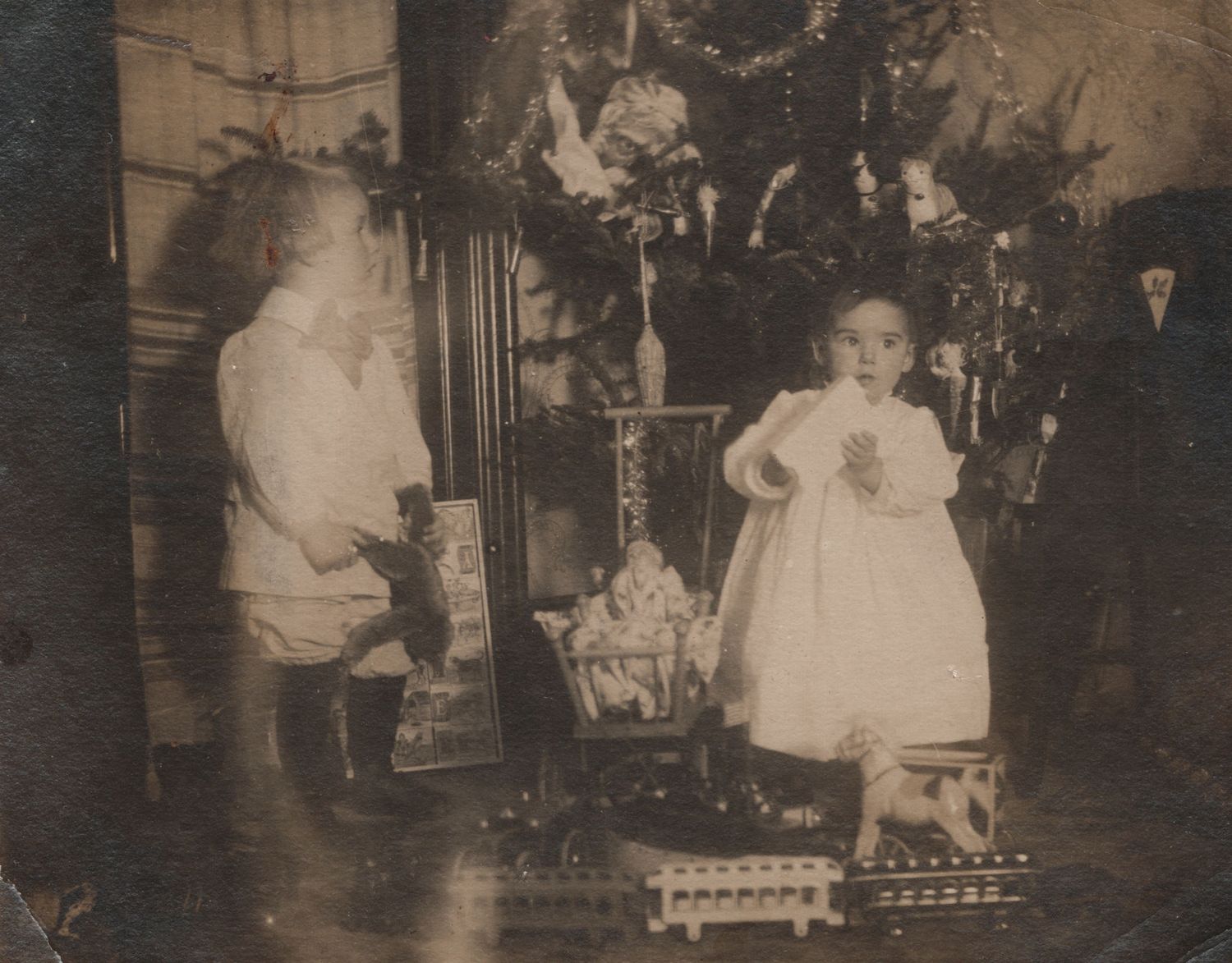 Vintage sepia photo of two young children in white clothing beside a decorated Christmas tree with toys.
