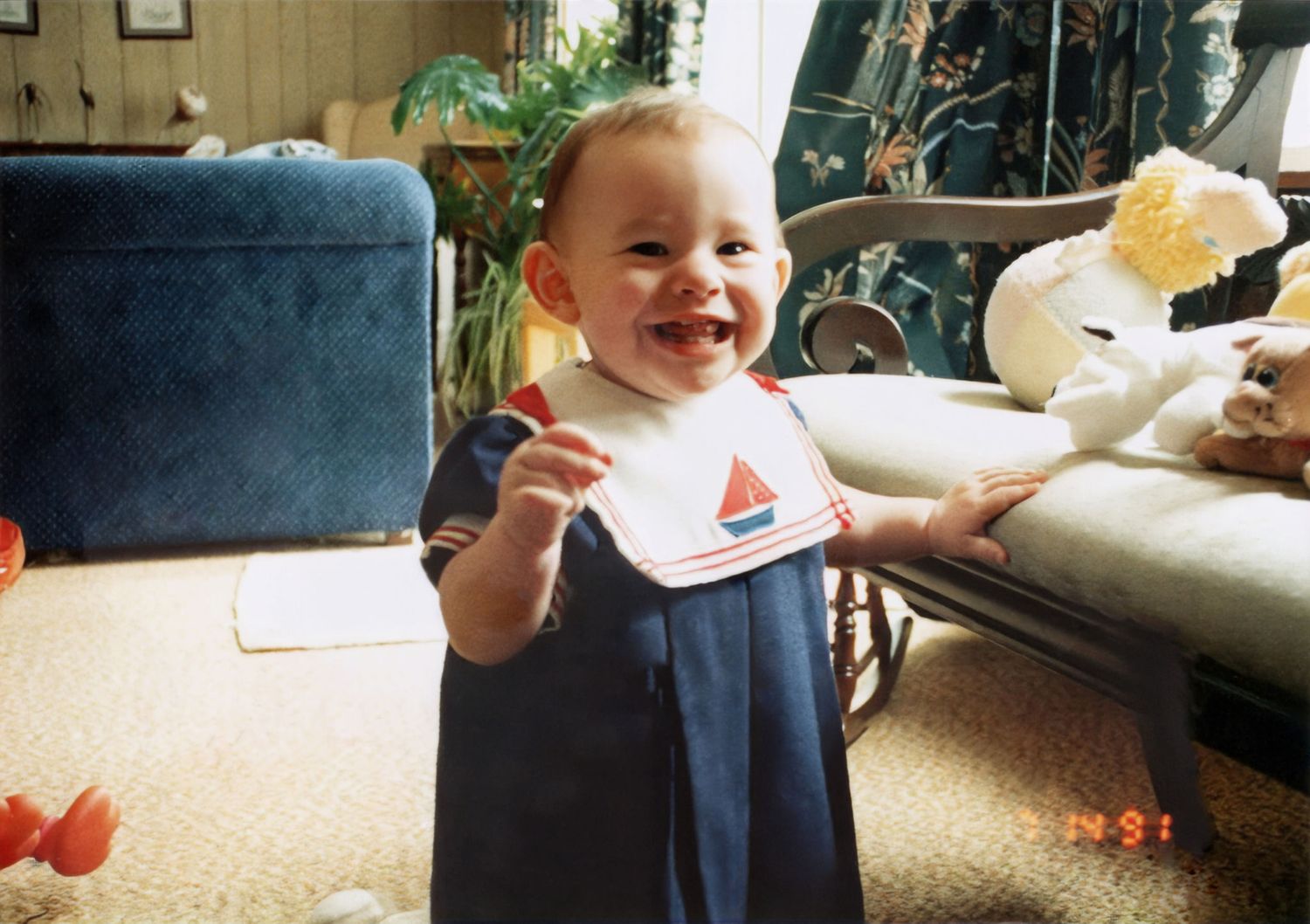Happy baby in navy sailor romper with bib, smiling and standing in a living room, vintage photo.