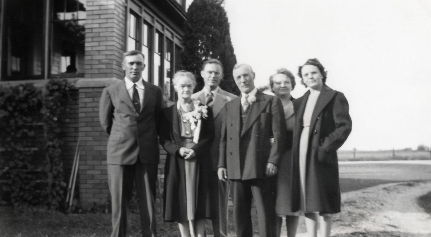 Vintage black and white family portrait of six adults posing outdoors beside a brick building on a sunny day.