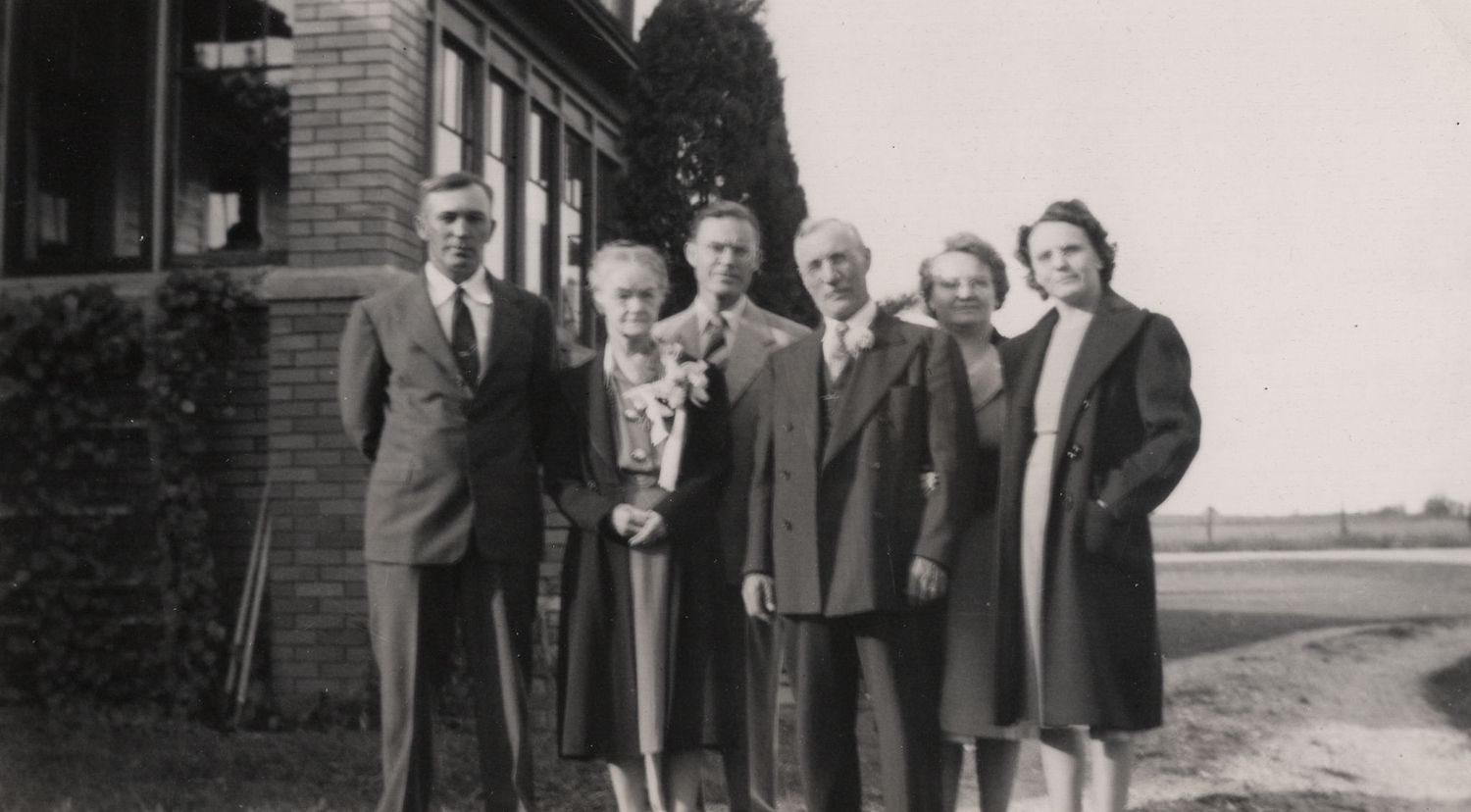 Vintage black and white photo of six people posing outdoors near a brick building in formal attire.