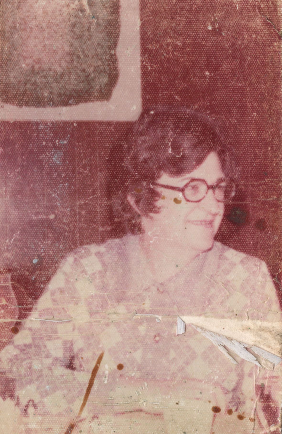 Vintage faded photo of a woman with round glasses and short dark hair, smiling softly in a patterned blouse.