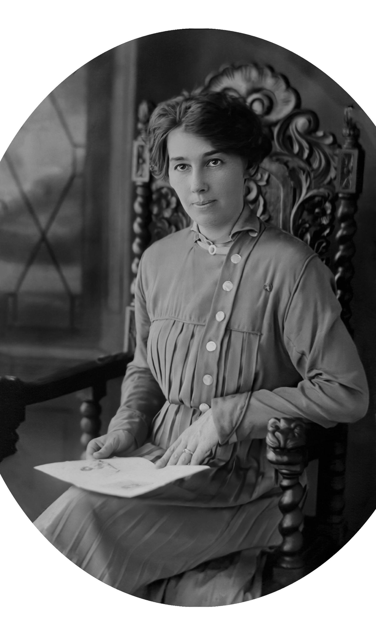 Vintage black and white oval portrait of a young woman in Victorian dress seated in an ornate wooden chair holding papers.
