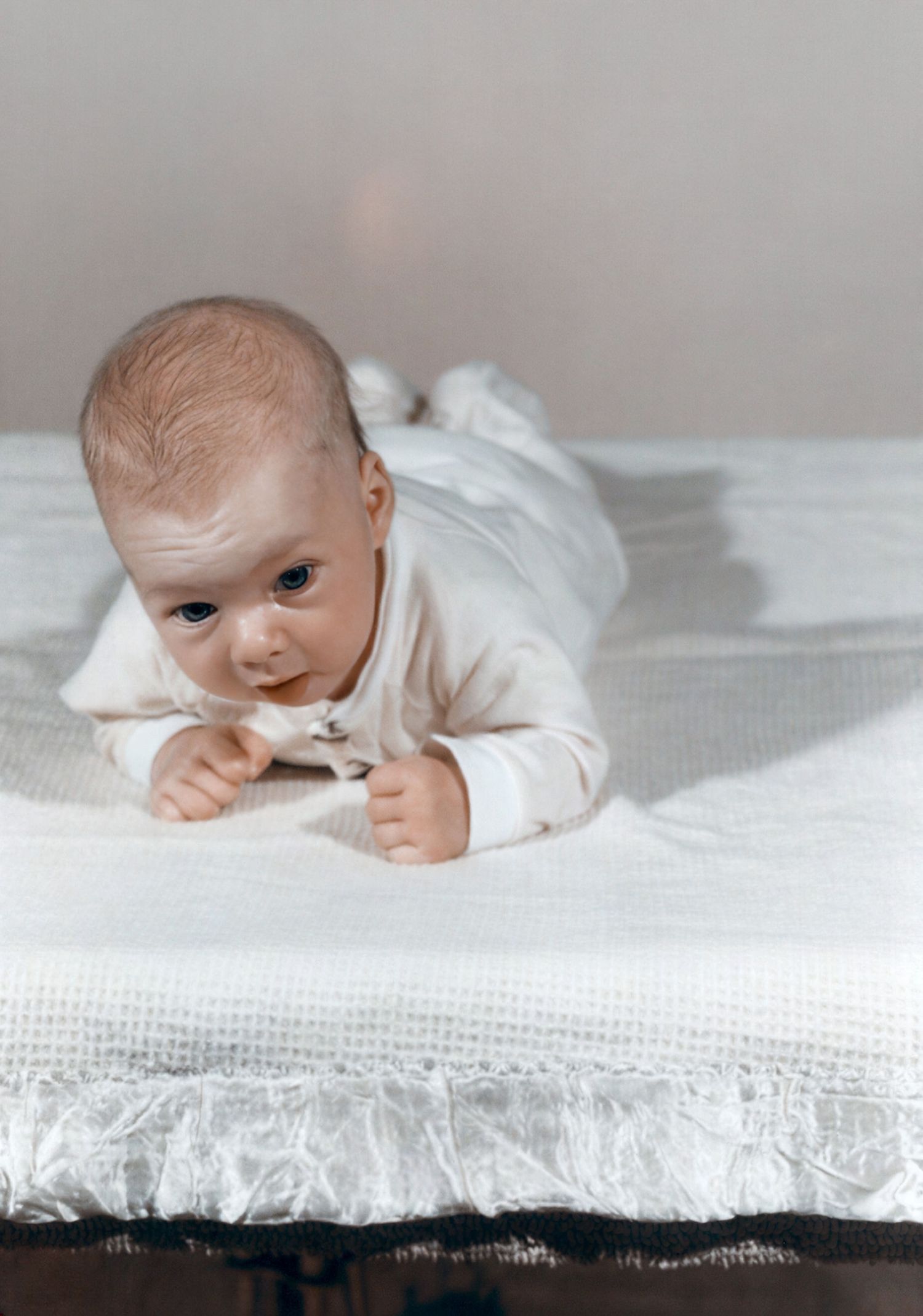 A newborn baby in a white onesie doing tummy time on a soft white blanket, lifting their head up.
