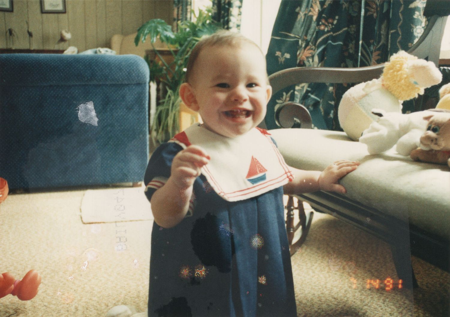 A happy baby in a navy blue sailor outfit with a bib smiles while standing in a living room.