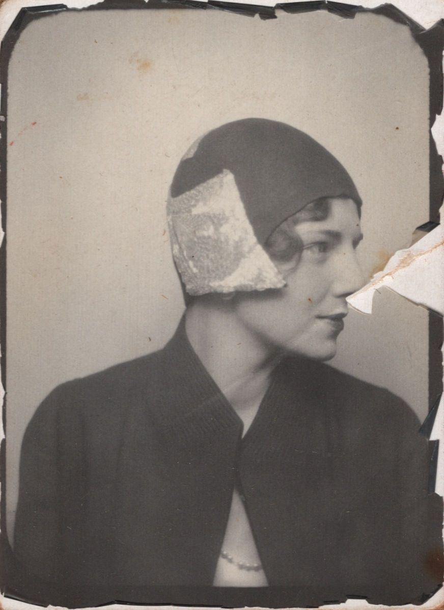 Vintage black and white photo booth portrait of a woman wearing a cloche hat with lace detail and dark jacket, 1920s style.