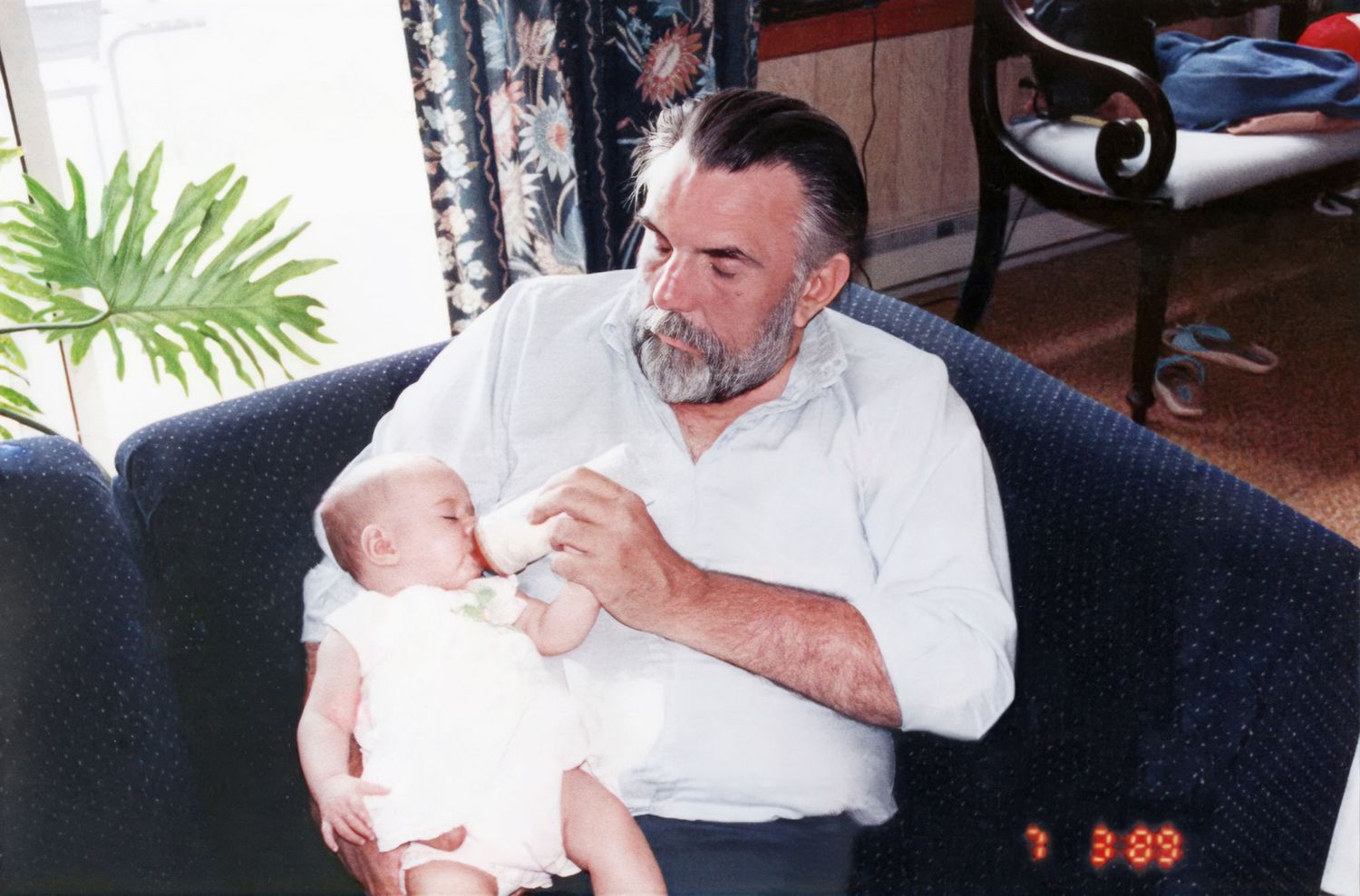 Elderly bearded man bottle feeding a newborn baby while sitting in a blue armchair.
