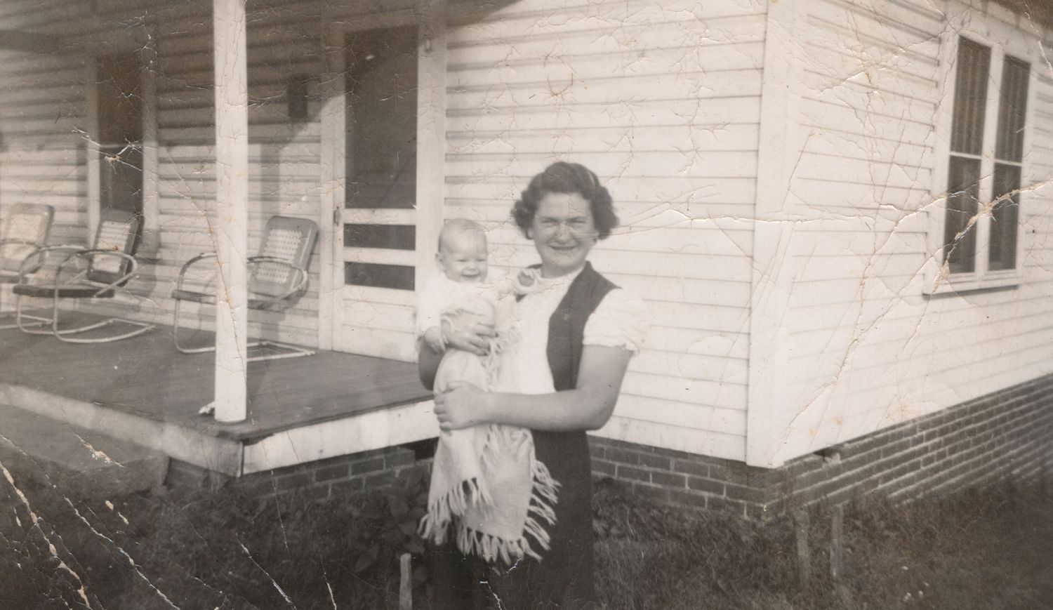 Vintage black and white photo of a woman holding a baby outside a white clapboard house with a porch.