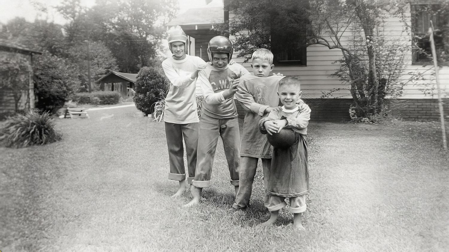 Vintage black and white photo of four children posing outdoors near a house surrounded by trees.