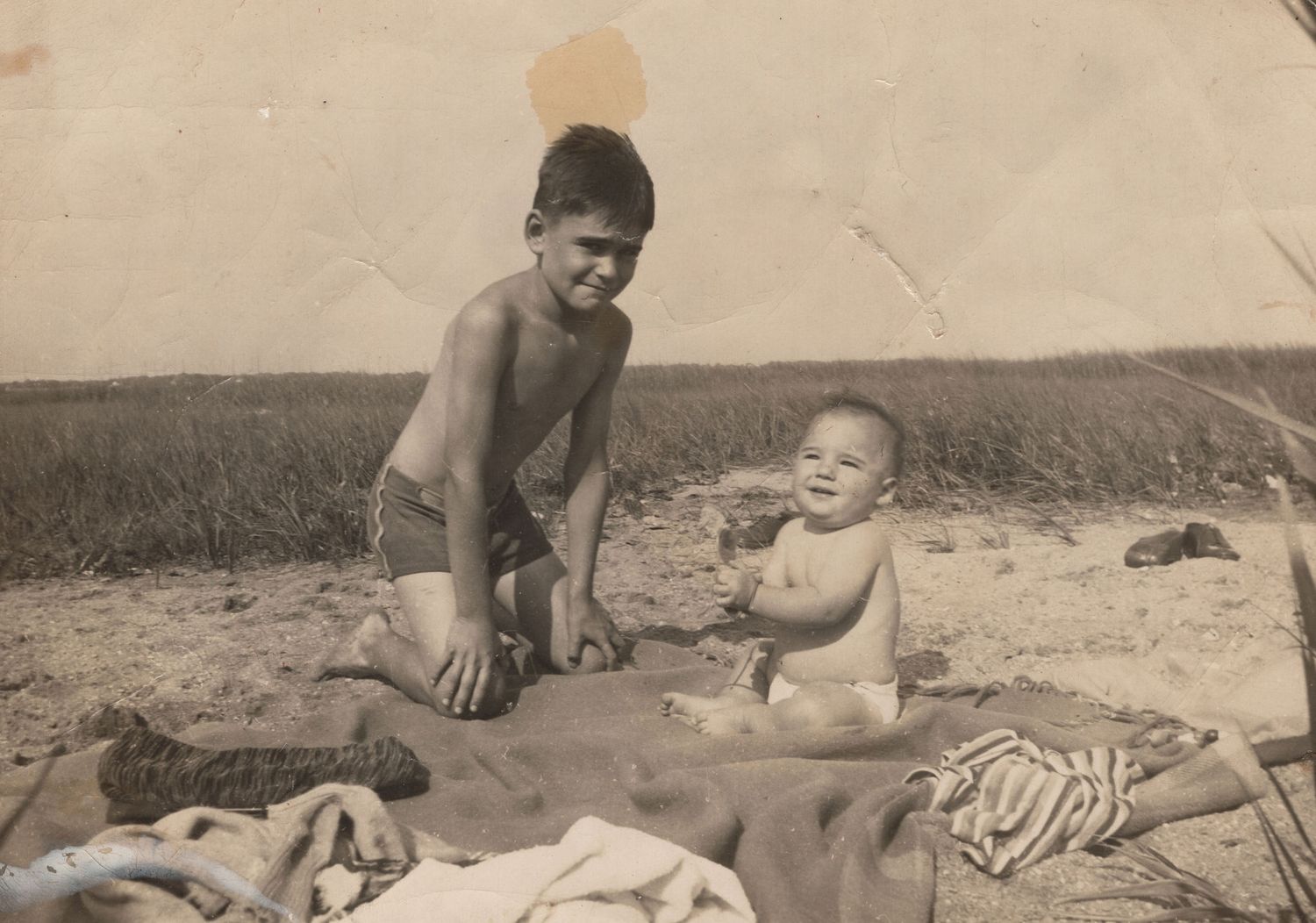 Vintage black and white photo of a young boy and baby sitting on a blanket at a sandy beach.